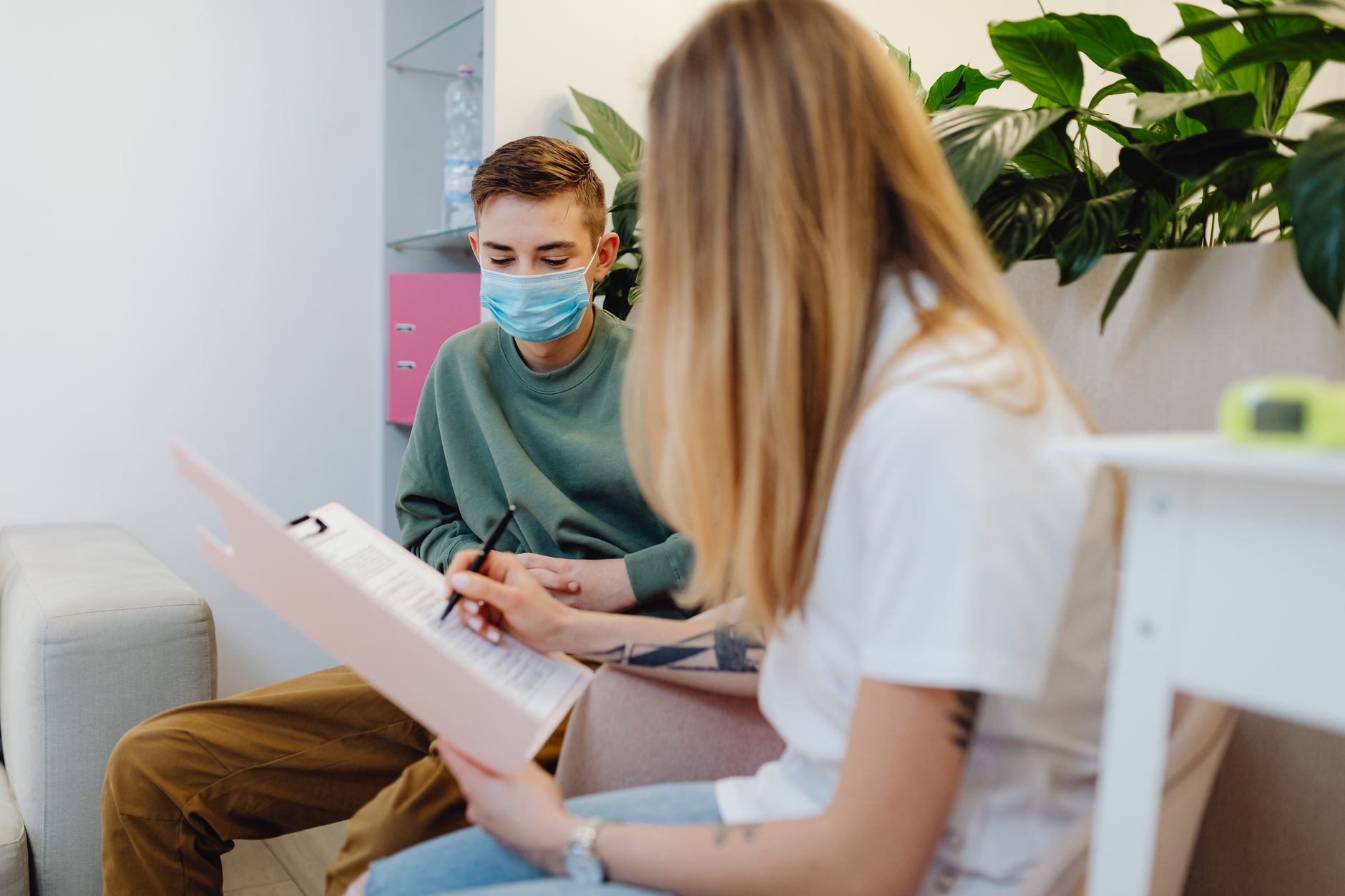 A psychologist in a session with a masked adolescent in a cozy clinic.