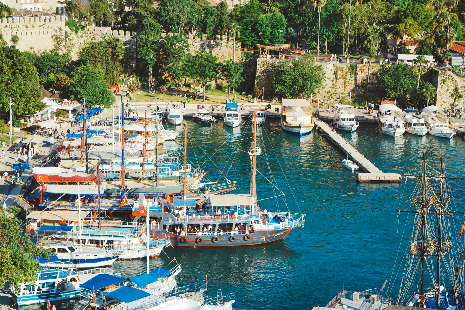A vibrant scene of boats docked at Antalya's old harbour during daylight, with lush greenery in the background.