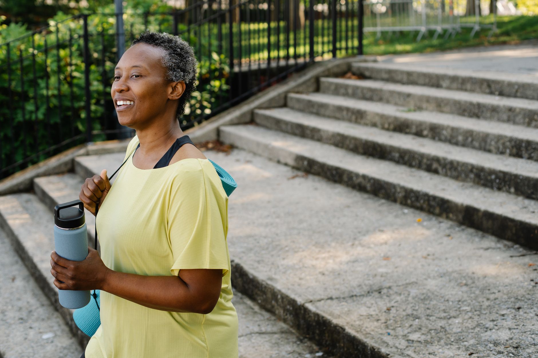 Smiling woman in yellow shirt holding a water bottle near stairs in a park.