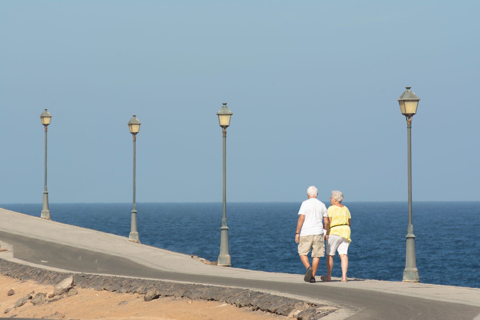 A senior couple strolls along a waterfront path in Izmir, with ocean views and lamp posts lining the way.