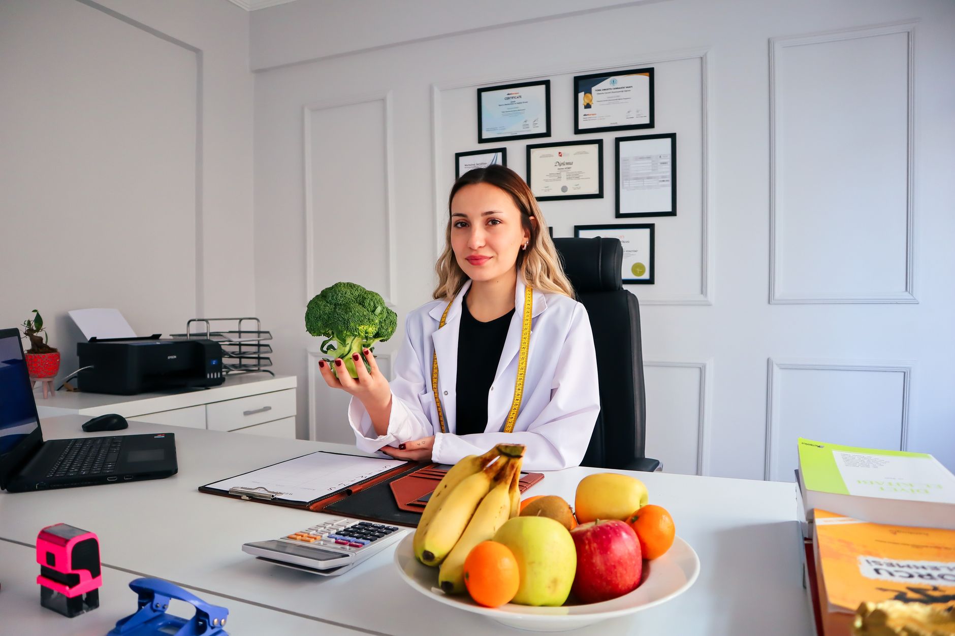 Dietitian holding broccoli in a consulting room with fruits on desk.