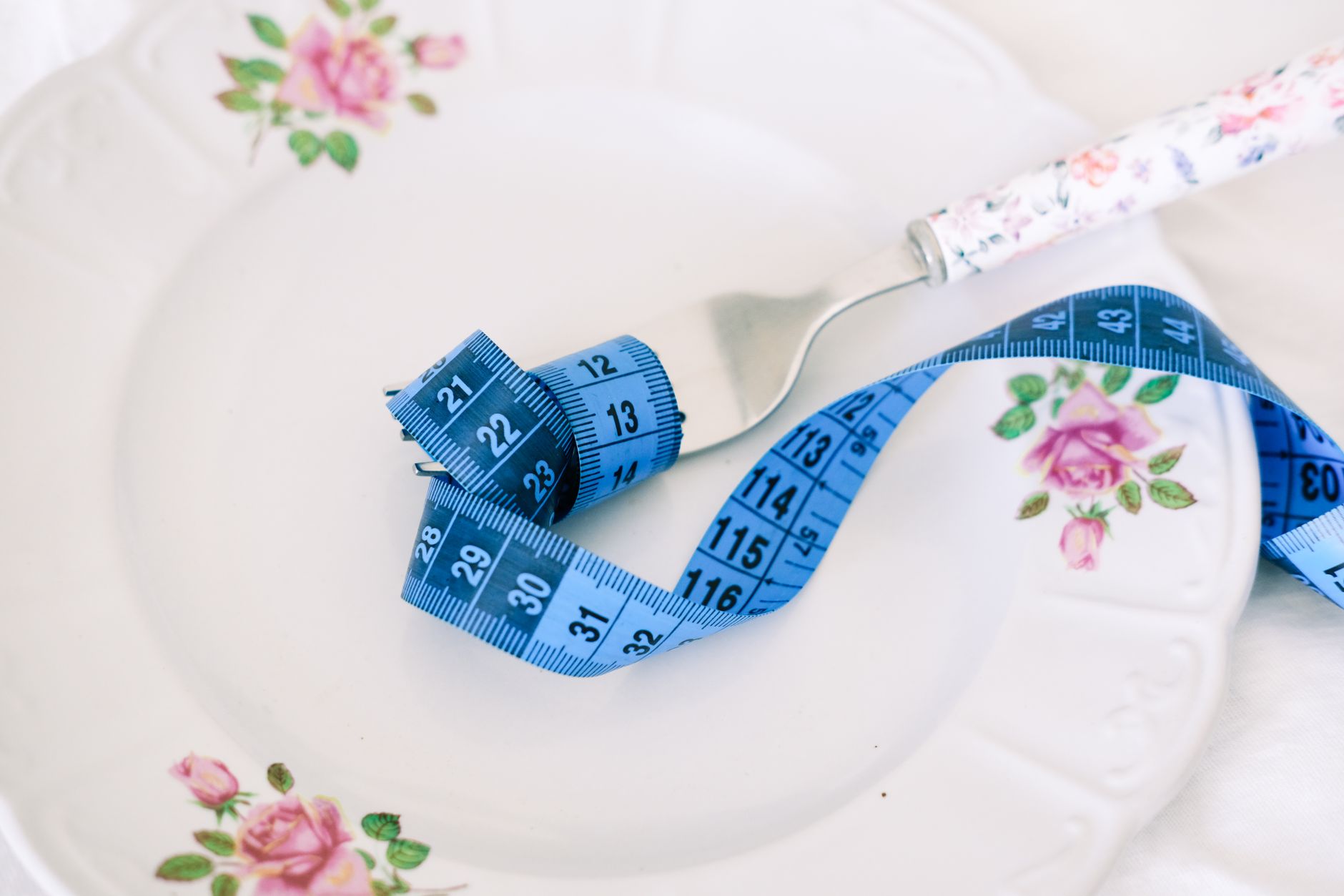 Blue measuring tape curled on a floral plate with a fork, symbolizing portion control for post-surgery nutrition.