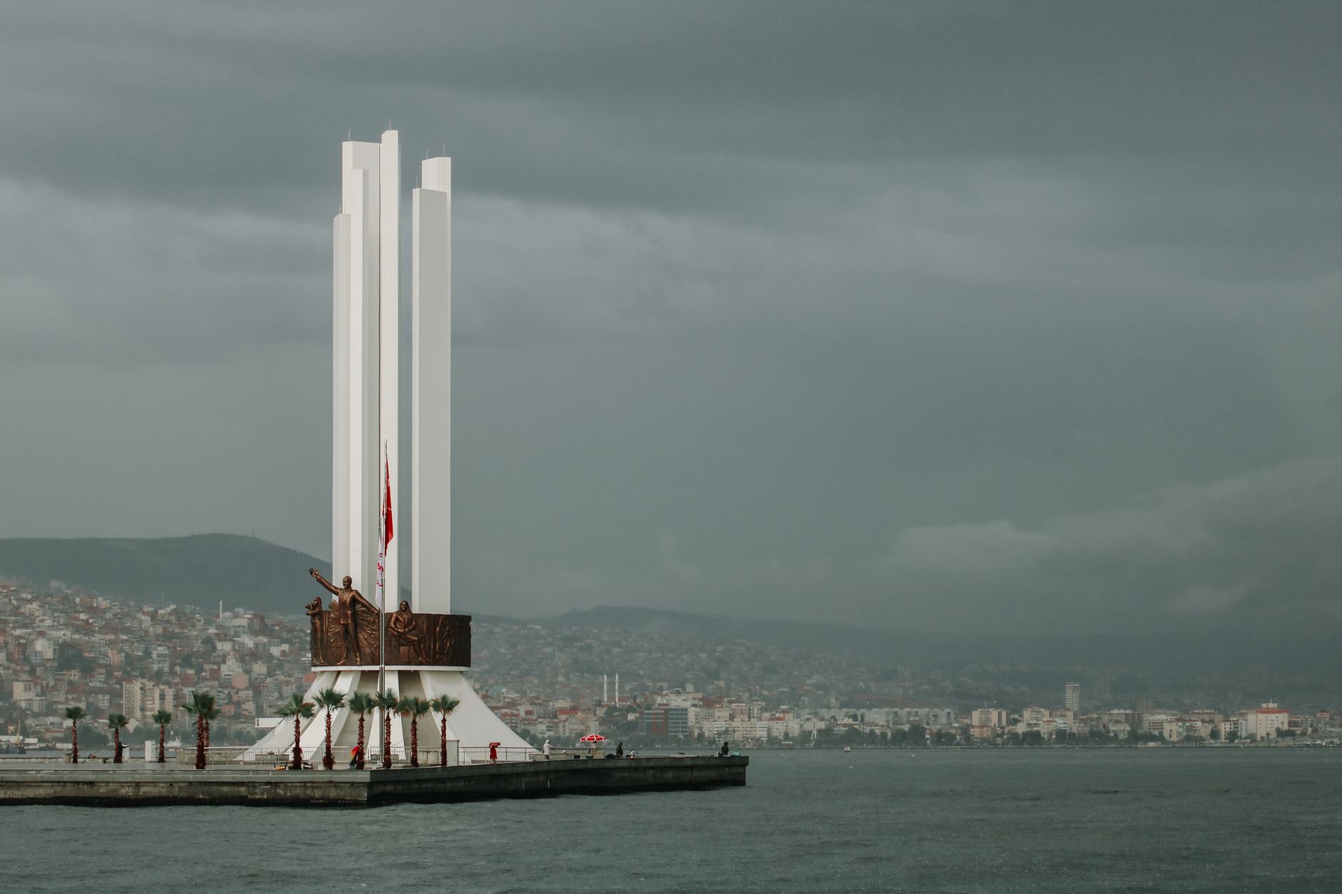 View of Izmir's coastline with a prominent monument, cloudy sky, and city buildings in the background.