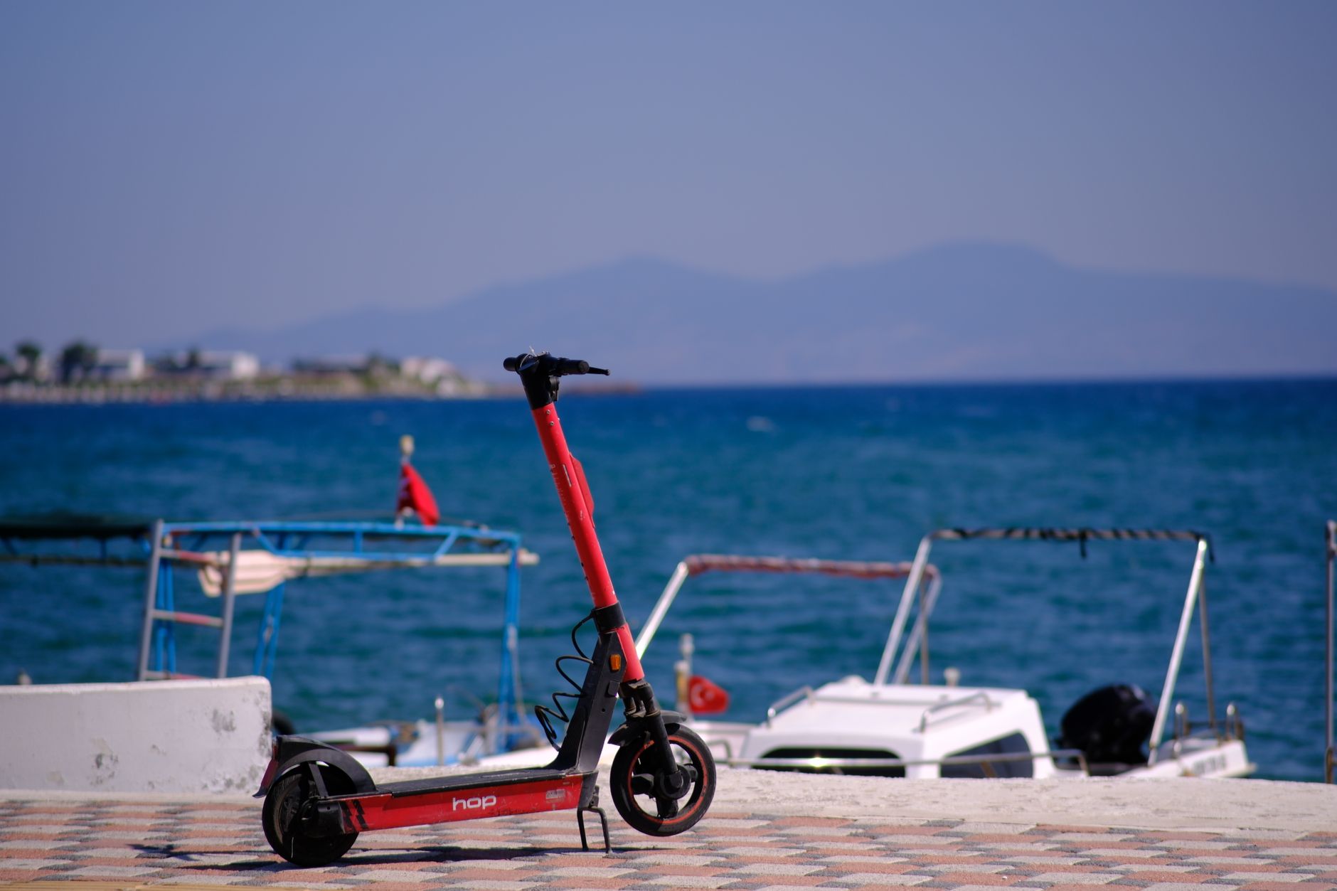 A red scooter parked near the harbor, with boats and sea in the background.