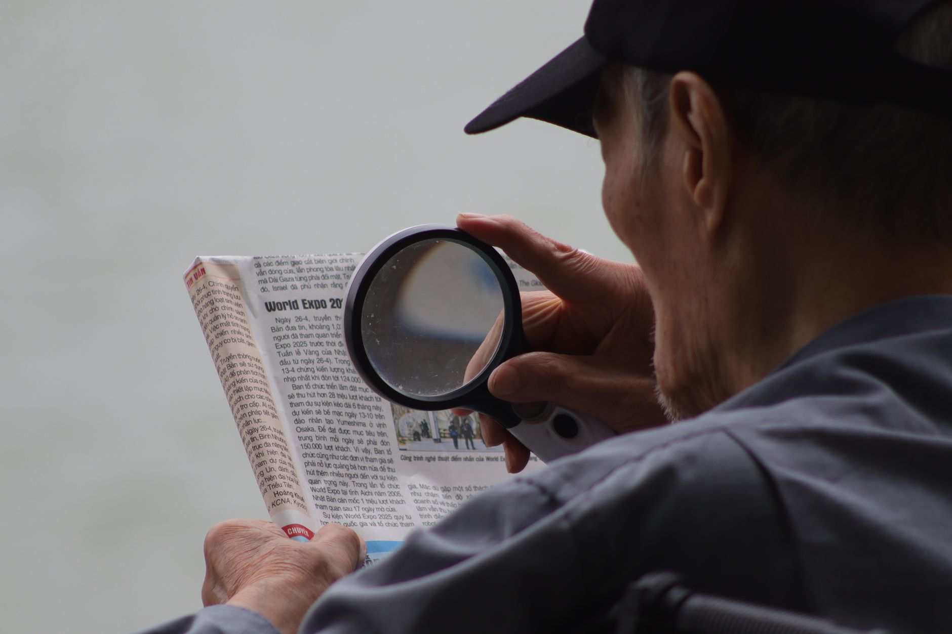 An elderly man uses a magnifying glass to read a newspaper by the water.