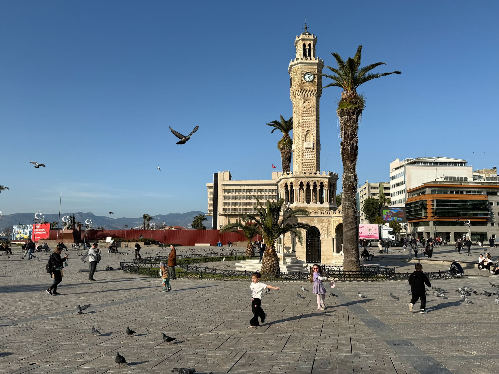 A sunny day in Izmir featuring the famous clock tower, people, and flying pigeons.