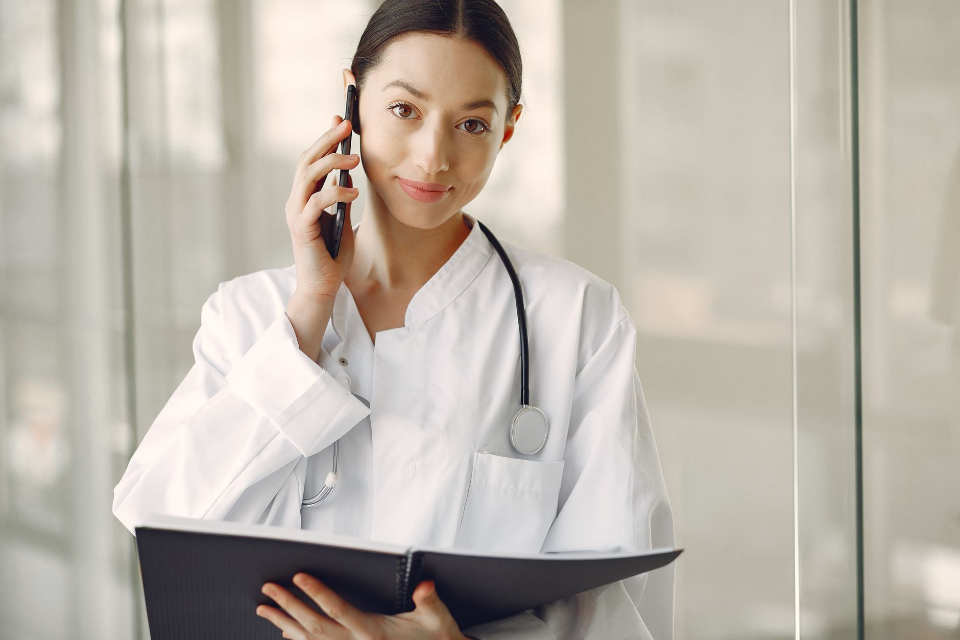 A doctor in a white coat speaking on the phone, holding a notebook, in a hospital setting.