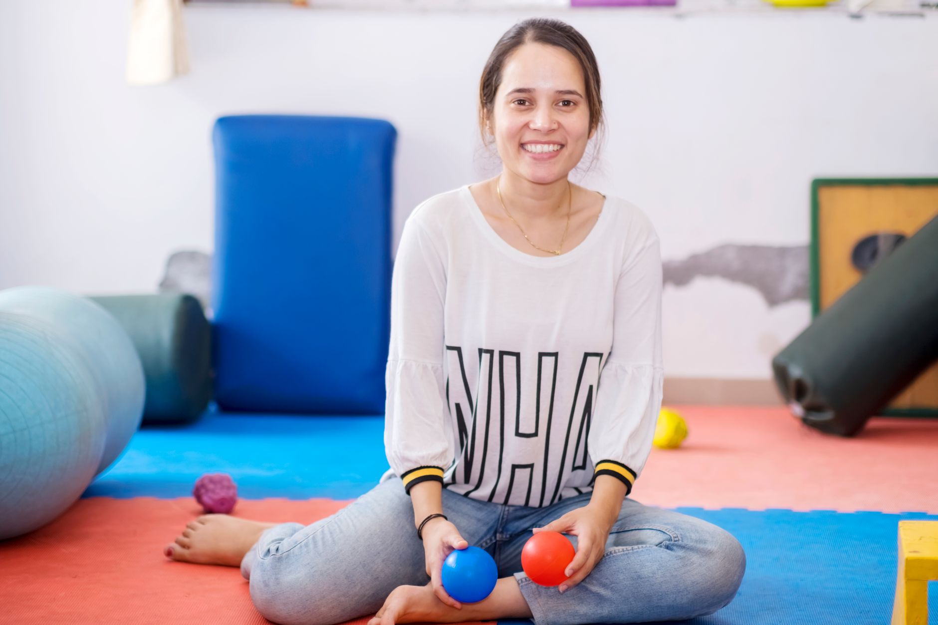 A speech-language therapist smiling while holding red and blue balls in a vibrant therapy room.