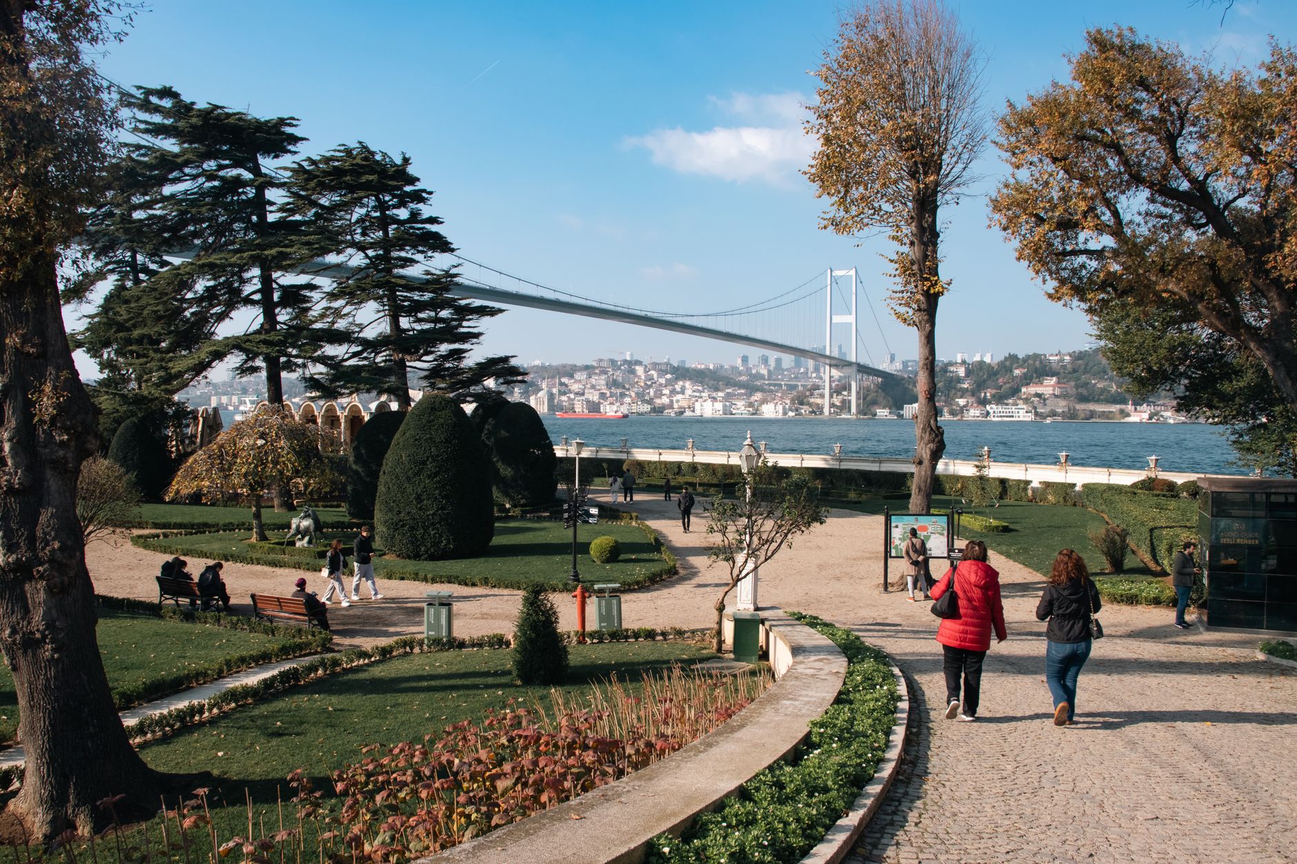 Two people stroll through a lush park in Istanbul with a view of the Bosphorus bridge.
