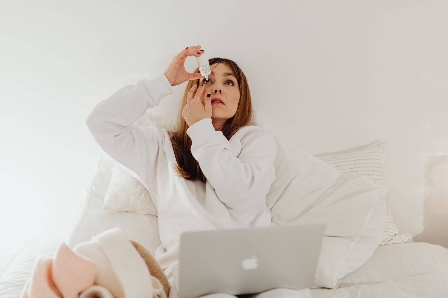 A patient in a white sweatshirt sitting on a bed, administering eye drops carefully.