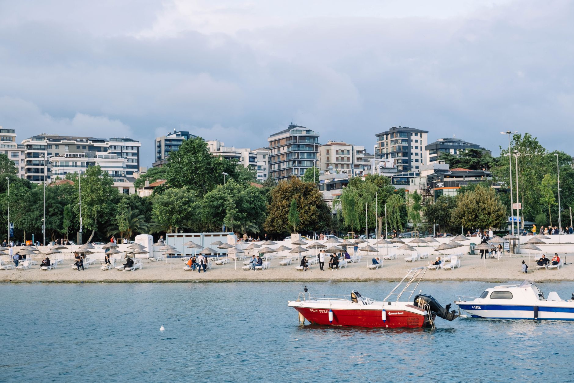 A coastal scene featuring two boats, sandy beach, modern buildings, and visitors enjoying the terrace.