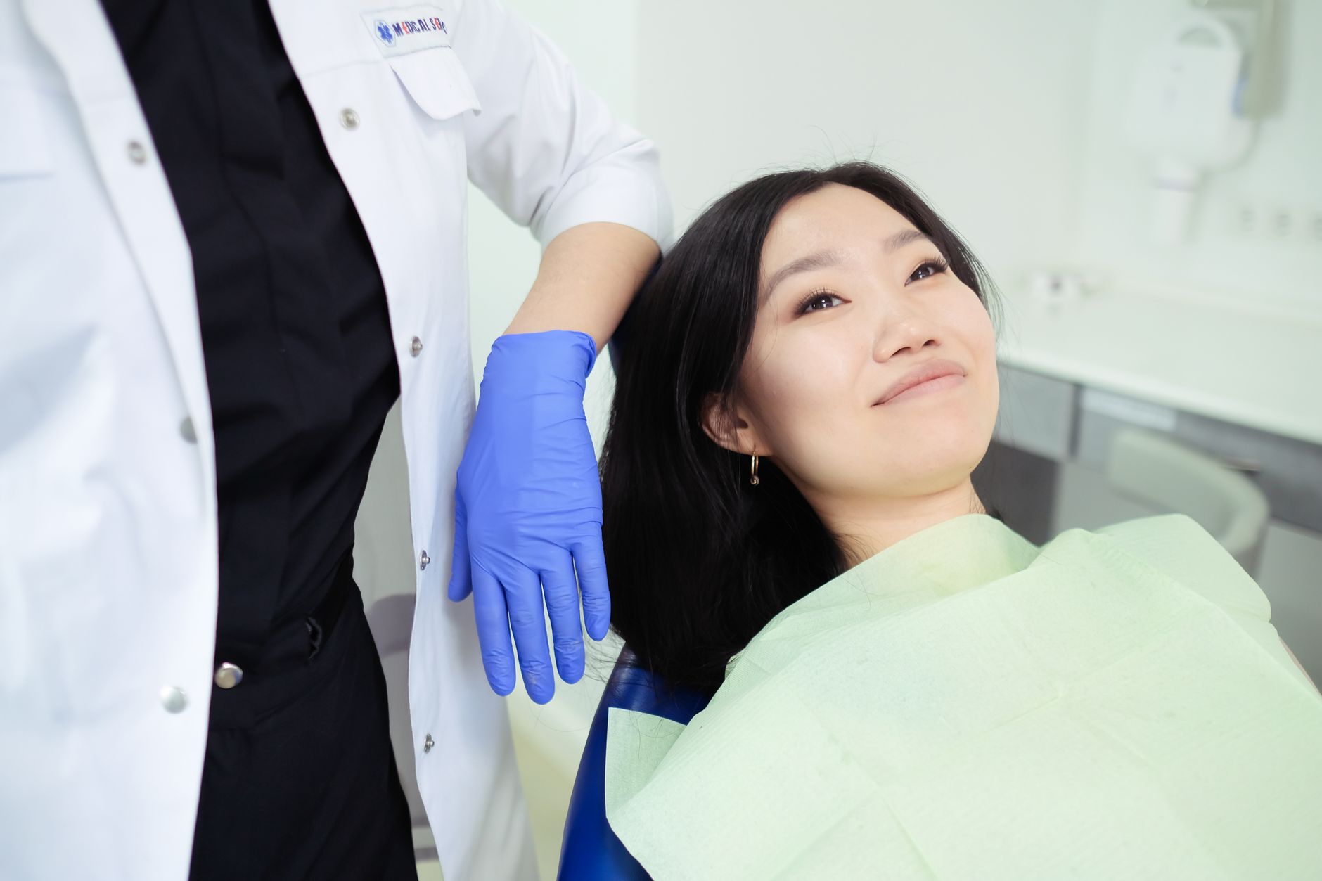 A smiling patient in a dental chair, wearing a green bib, with a dentist's hand nearby.