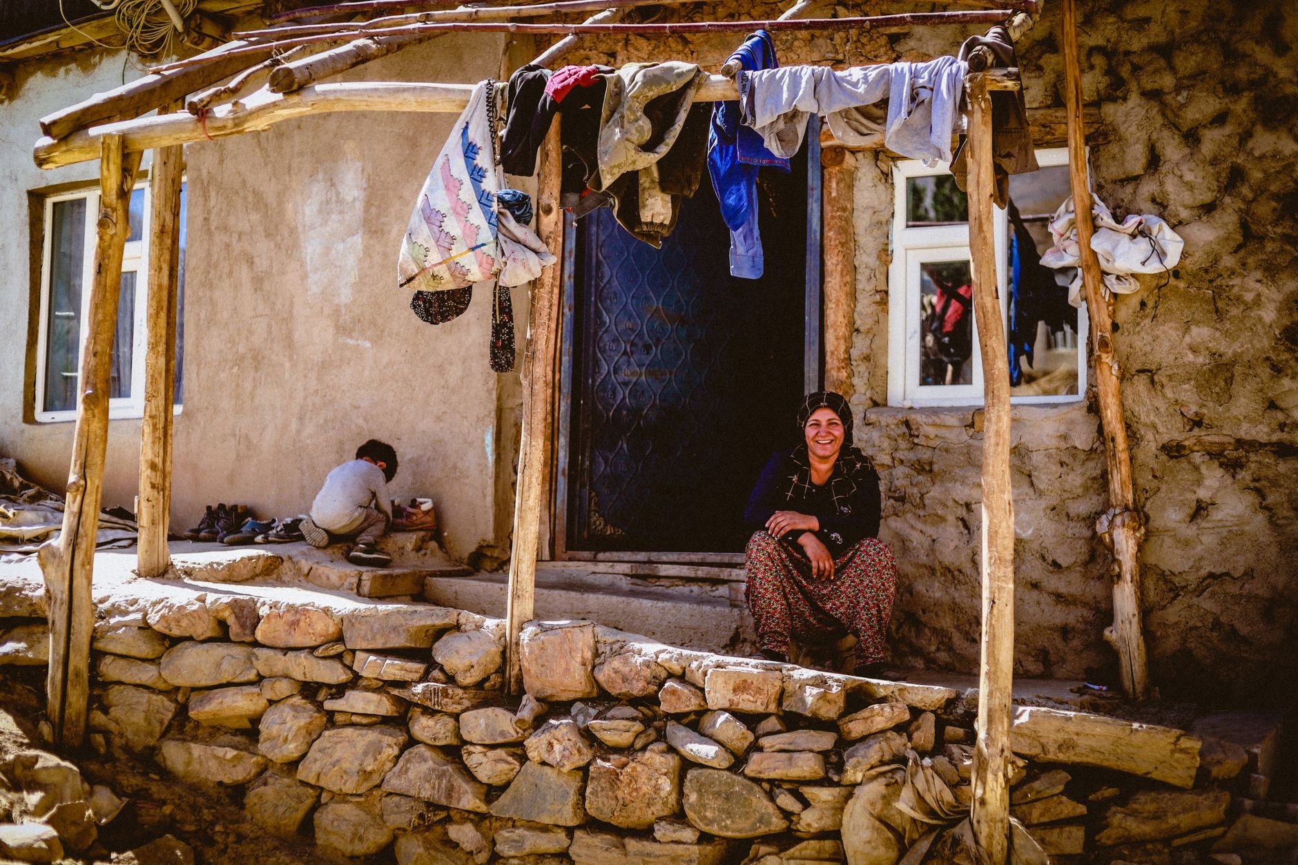A friendly woman sits on the porch of a village guesthouse with stone walls, while a child plays nearby.