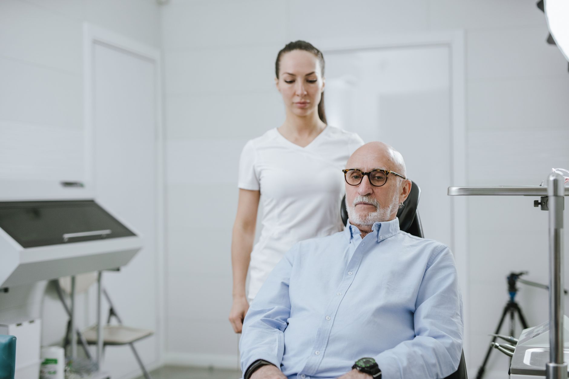 An older man seated in a clinic chair with glasses, looking serious, while a caregiver stands behind him.