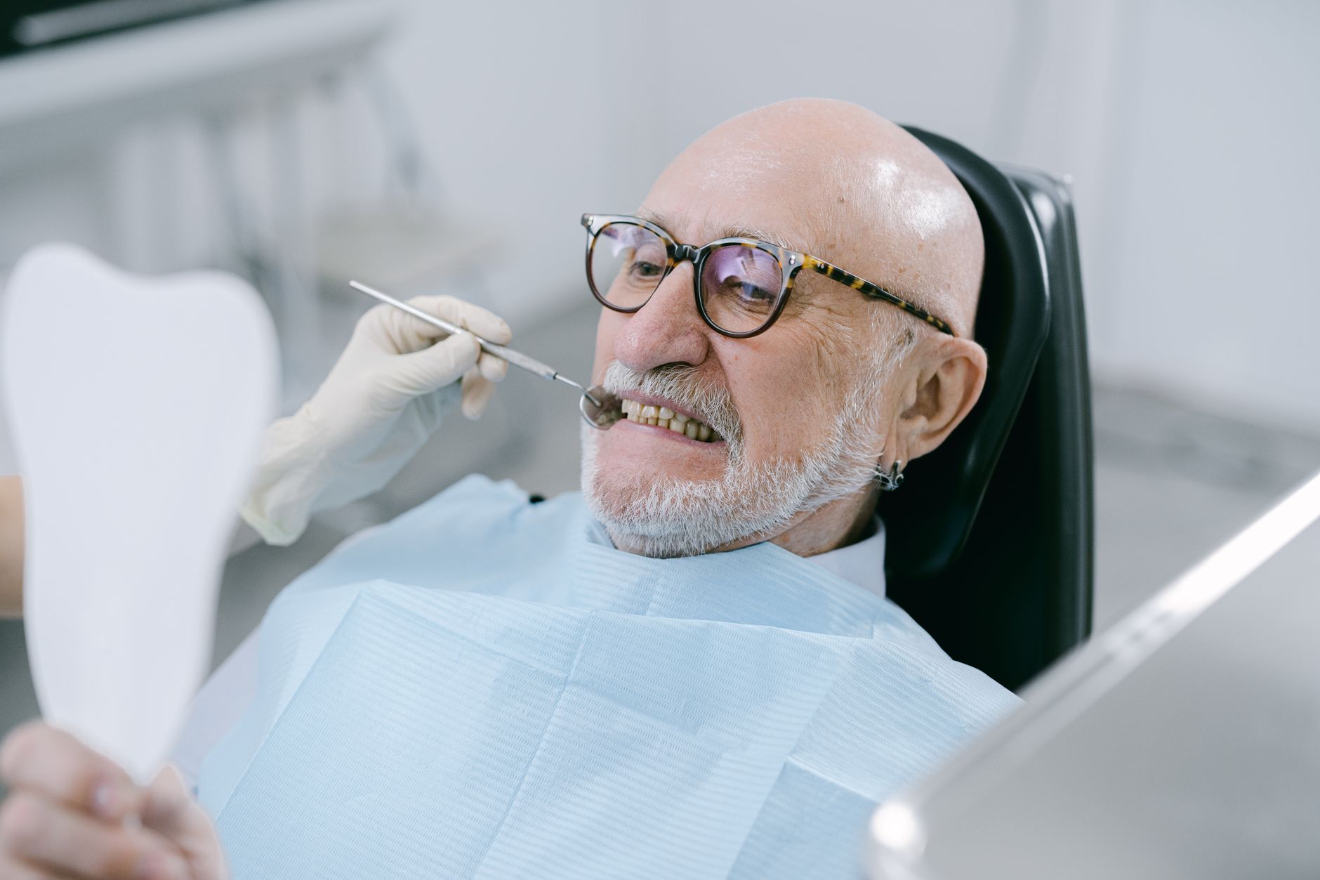 Elderly man smiling in dental chair during checkup, holding a mirror.