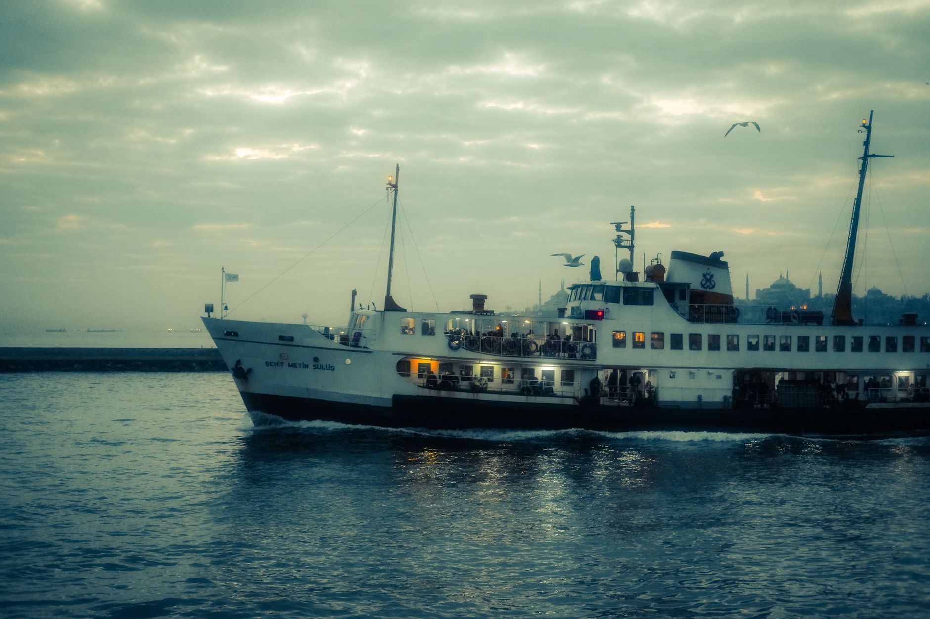 A ferry named 'Şehit Metin Sülü' sails through Istanbul's Bosphorus, surrounded by a tranquil evening sky.