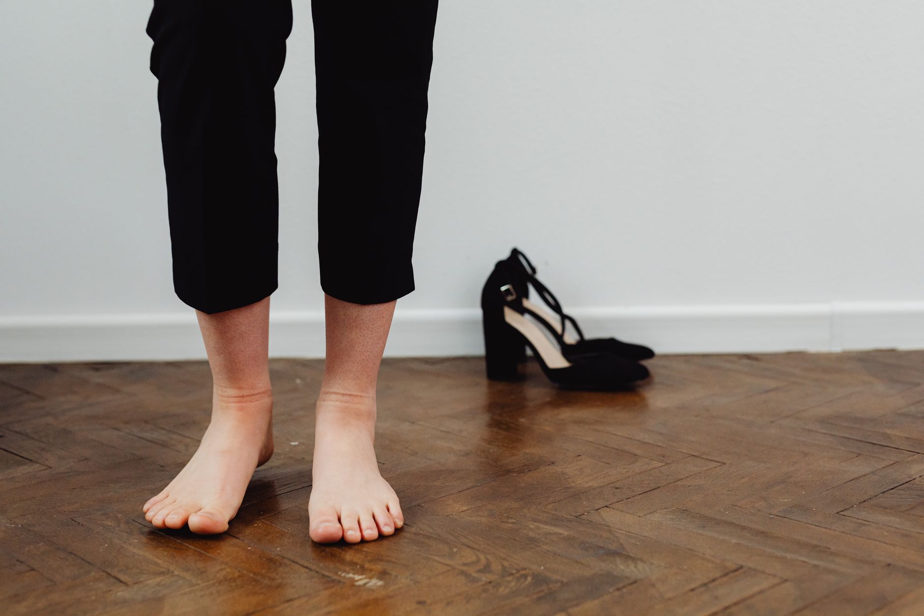 Feet standing barefoot on wooden flooring next to elegant black heels.