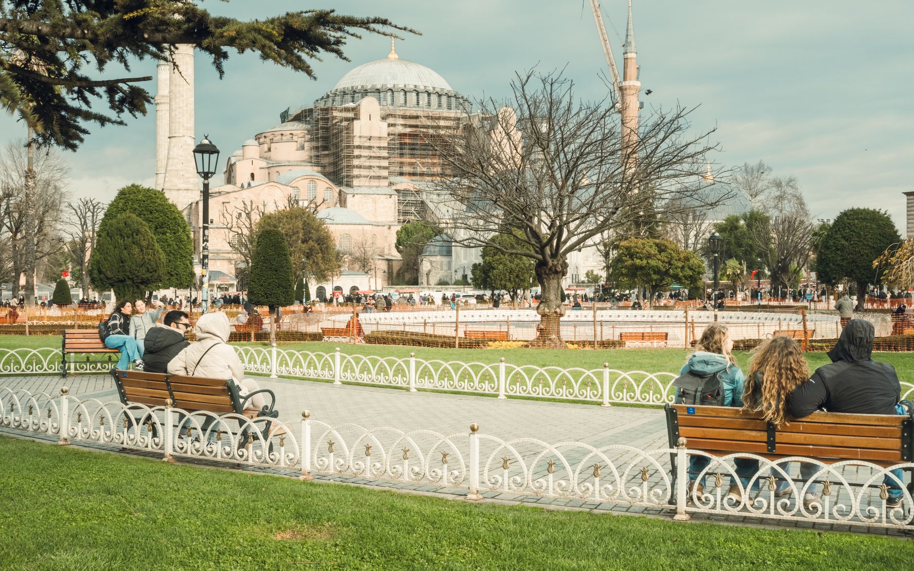 Visitors relax in a park with benches, overlooking the Hagia Sophia under renovation in Istanbul.