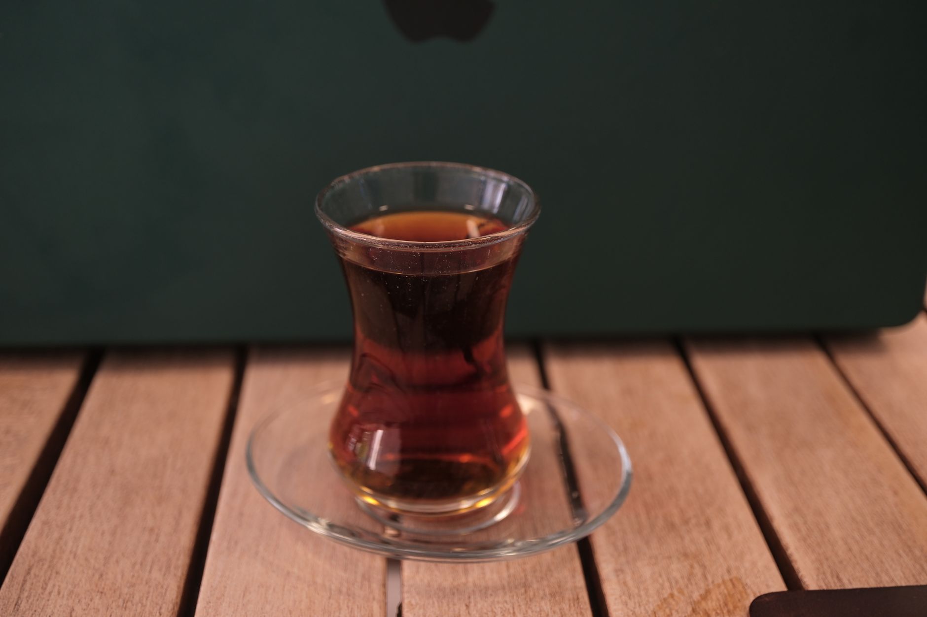 A glass of Turkish tea on a saucer, with a wooden table and a MacBook in the background.