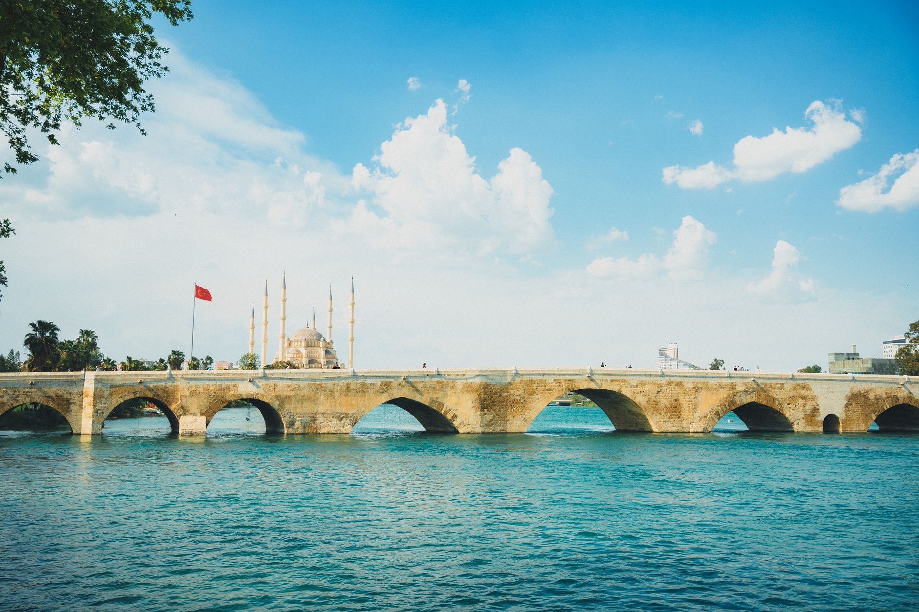 View of a historic bridge in Adana, Turkey, with a mosque and Turkish flag against a bright sky.
