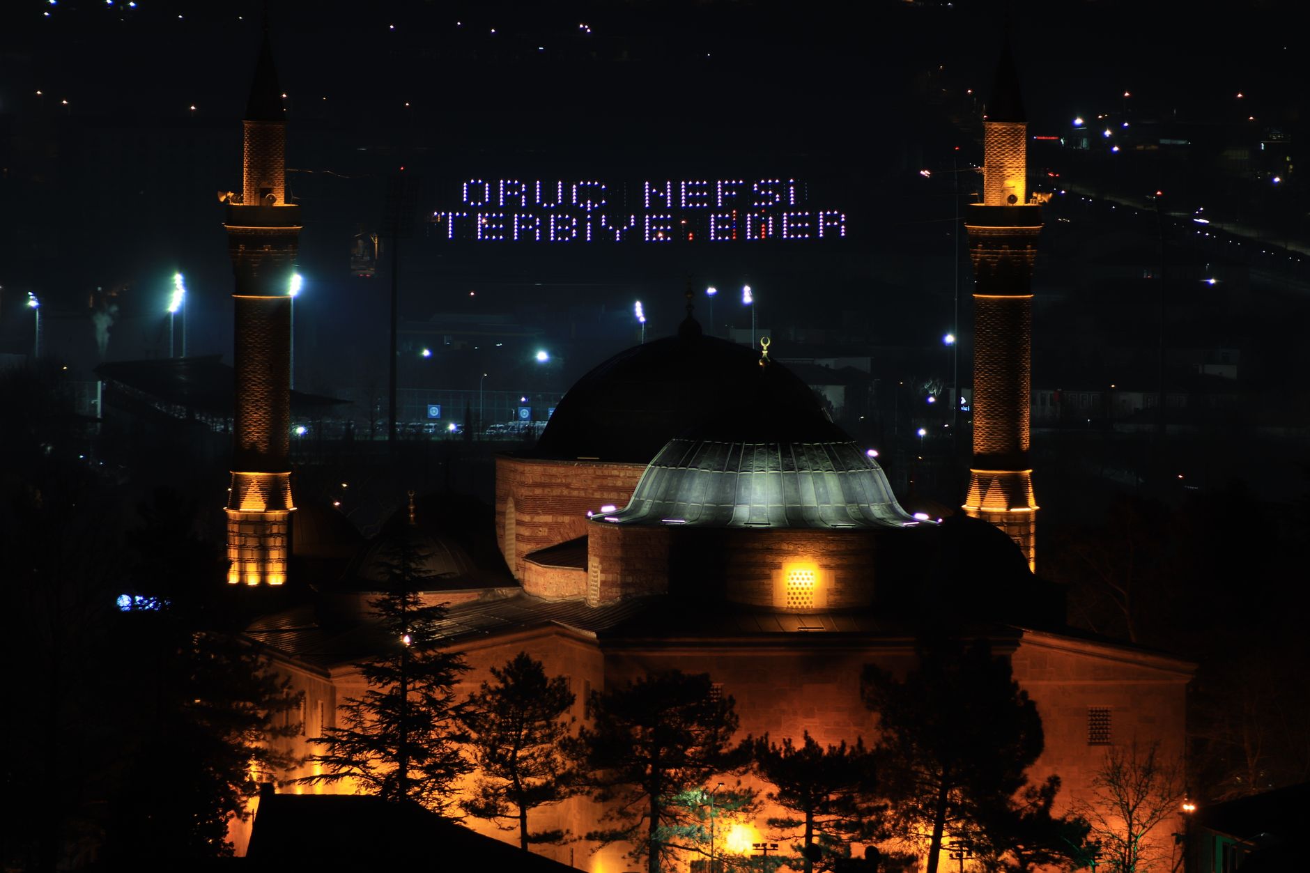 Night view of the Grand Mosque of Bursa, beautifully lit, with a digital sign reading 'Oruç Nefsi Terbiye Eder'.