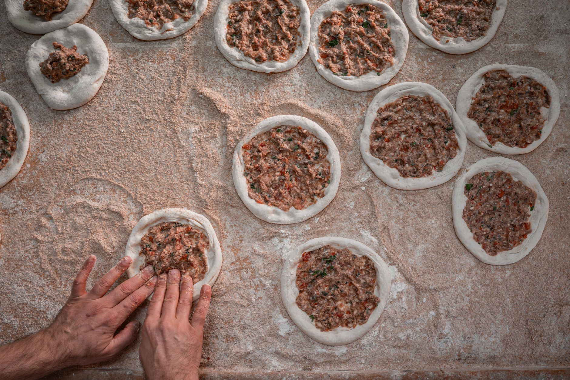 Unlanmış bir yüzeyde baharatlı kıyma harcıyla lahmacun şekillendiren eller.