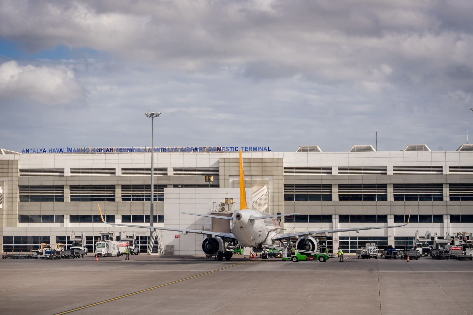 View of Antalya Airport Domestic Terminal with a parked aircraft and ground service vehicles.