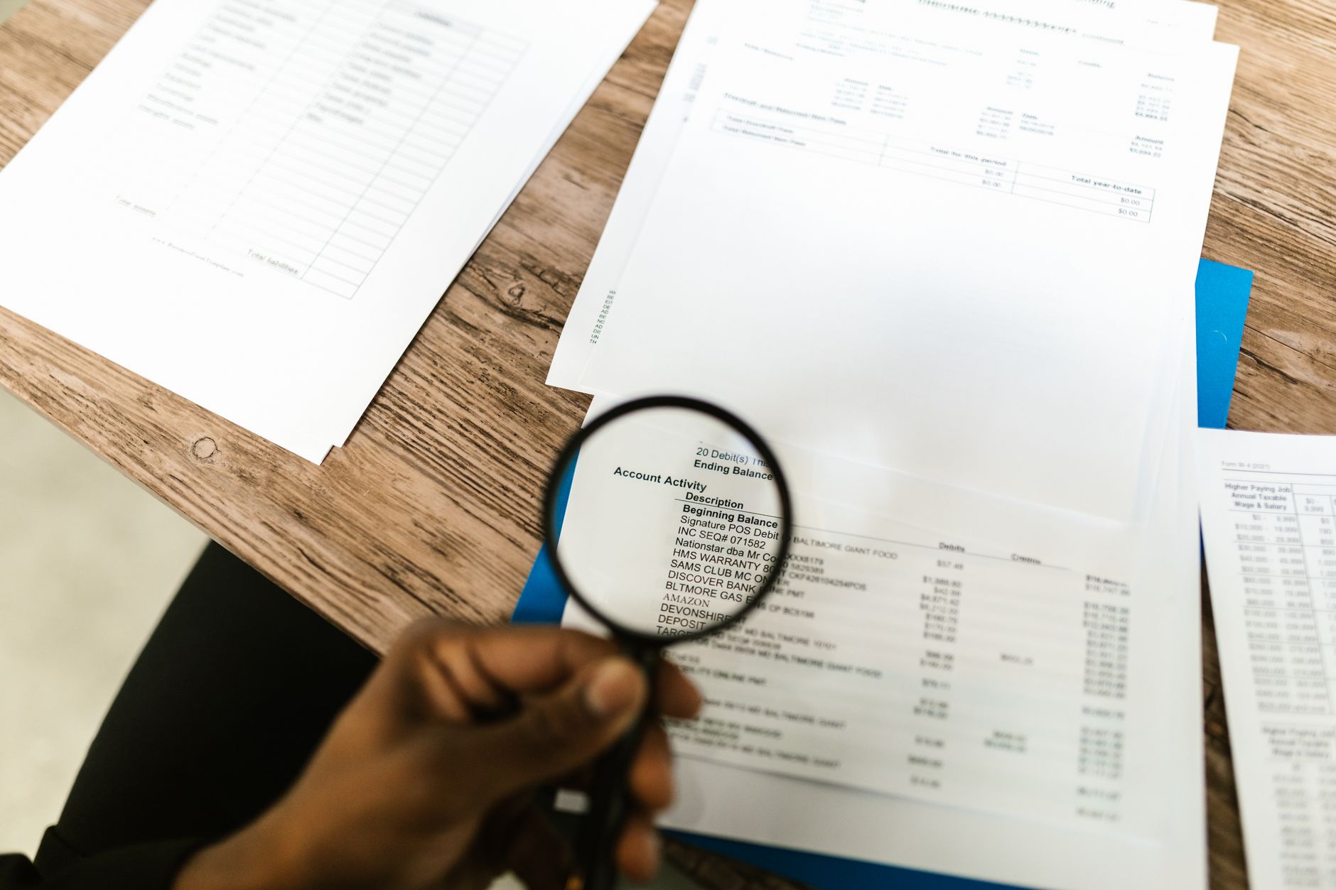 Close-up of a hand holding a magnifying glass over financial documents on a wooden table.