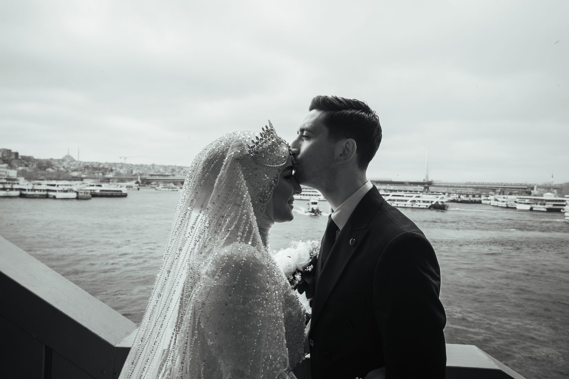 Bride and groom sharing a tender kiss on a dock in Türkiye, with boats and city skyline in the background.