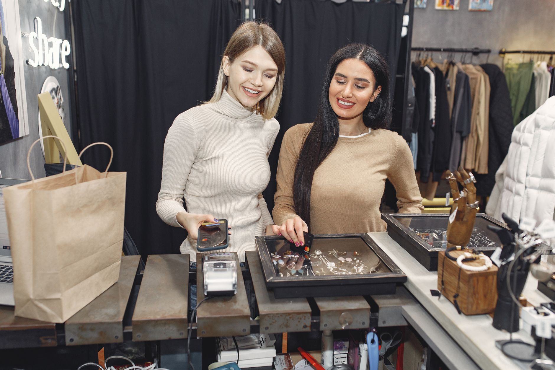 Two women smiling at a jewelry display, one holding a smartphone, in a stylish shop.
