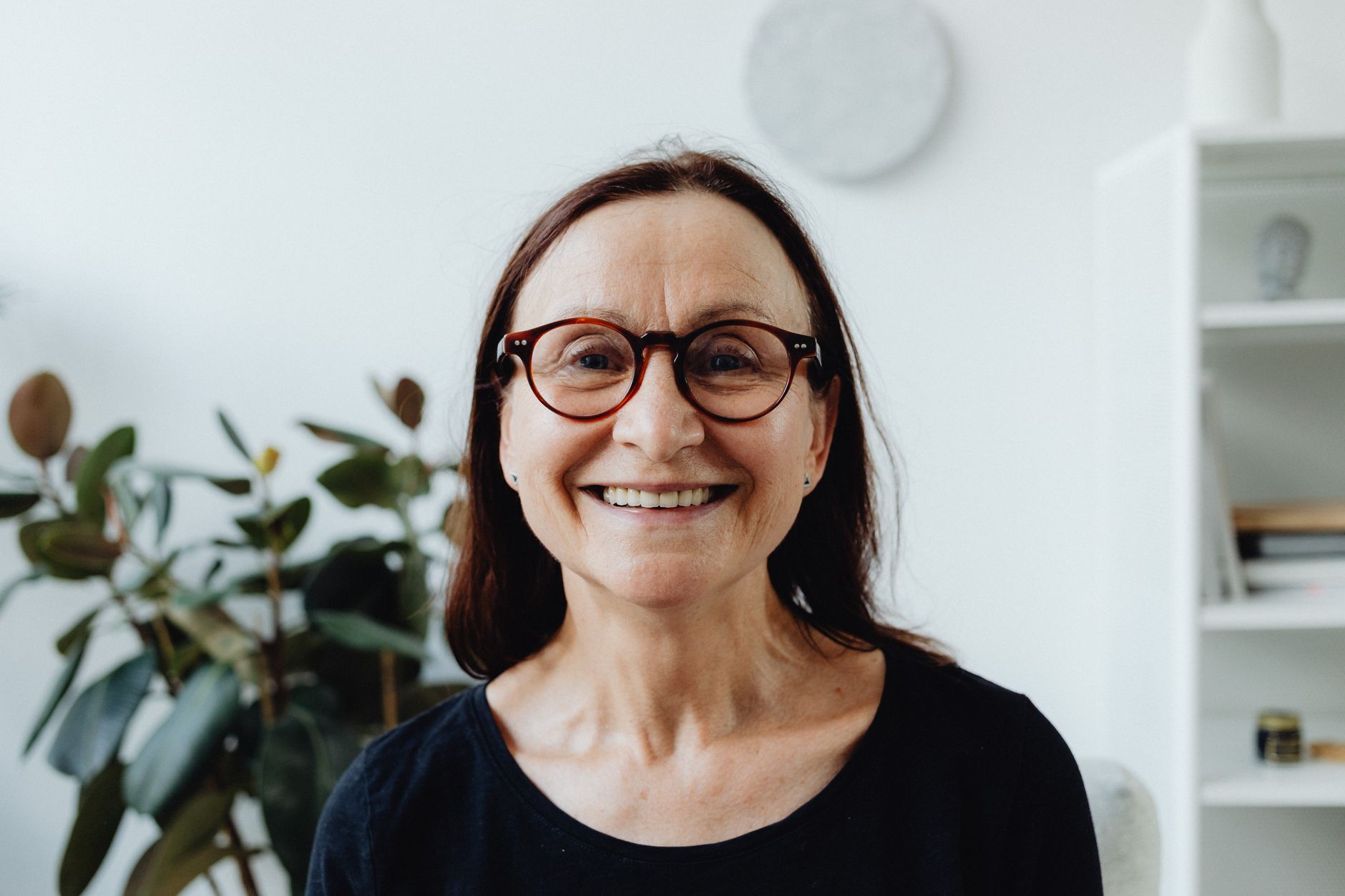A smiling woman with long hair and glasses, seated in a bright, modern space.