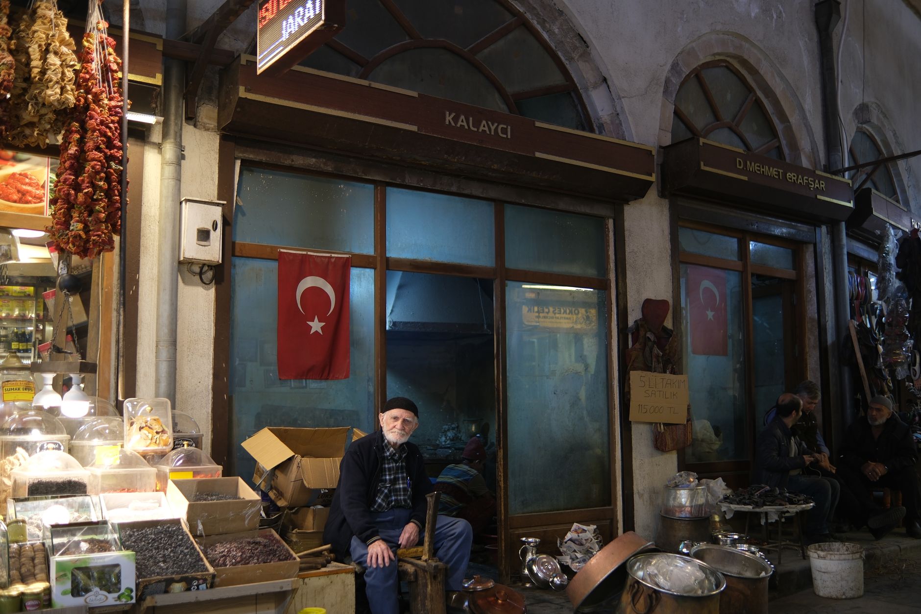 Yaşlı bir dükkân sahibi İstanbul çarşısında dükkânının önünde oturuyor, etrafı baharatlar ve mallarla çevrili, sıcak bir gülümseme sunuyor.