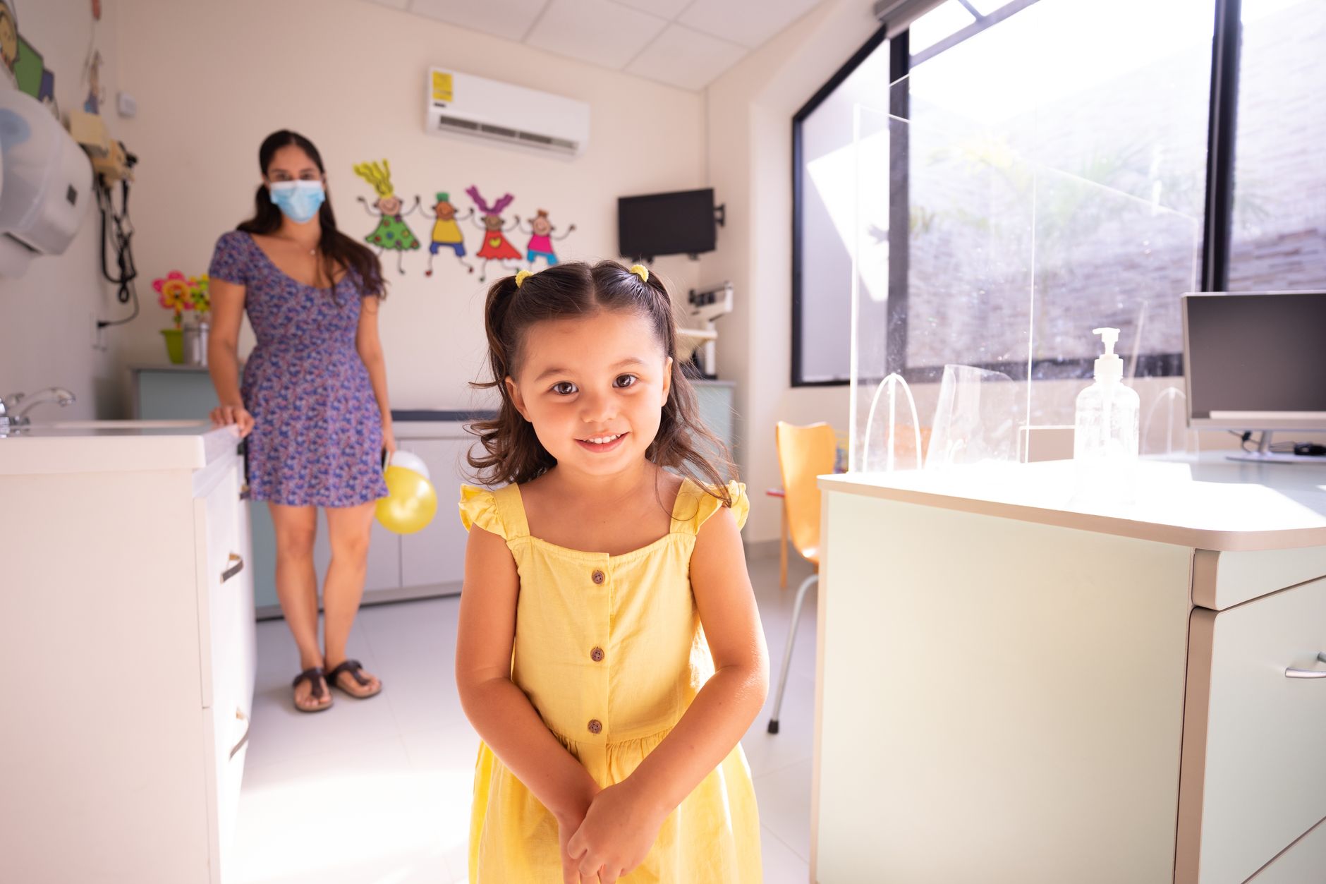 A smiling girl in a yellow dress stands in a colorful children's hospital room.