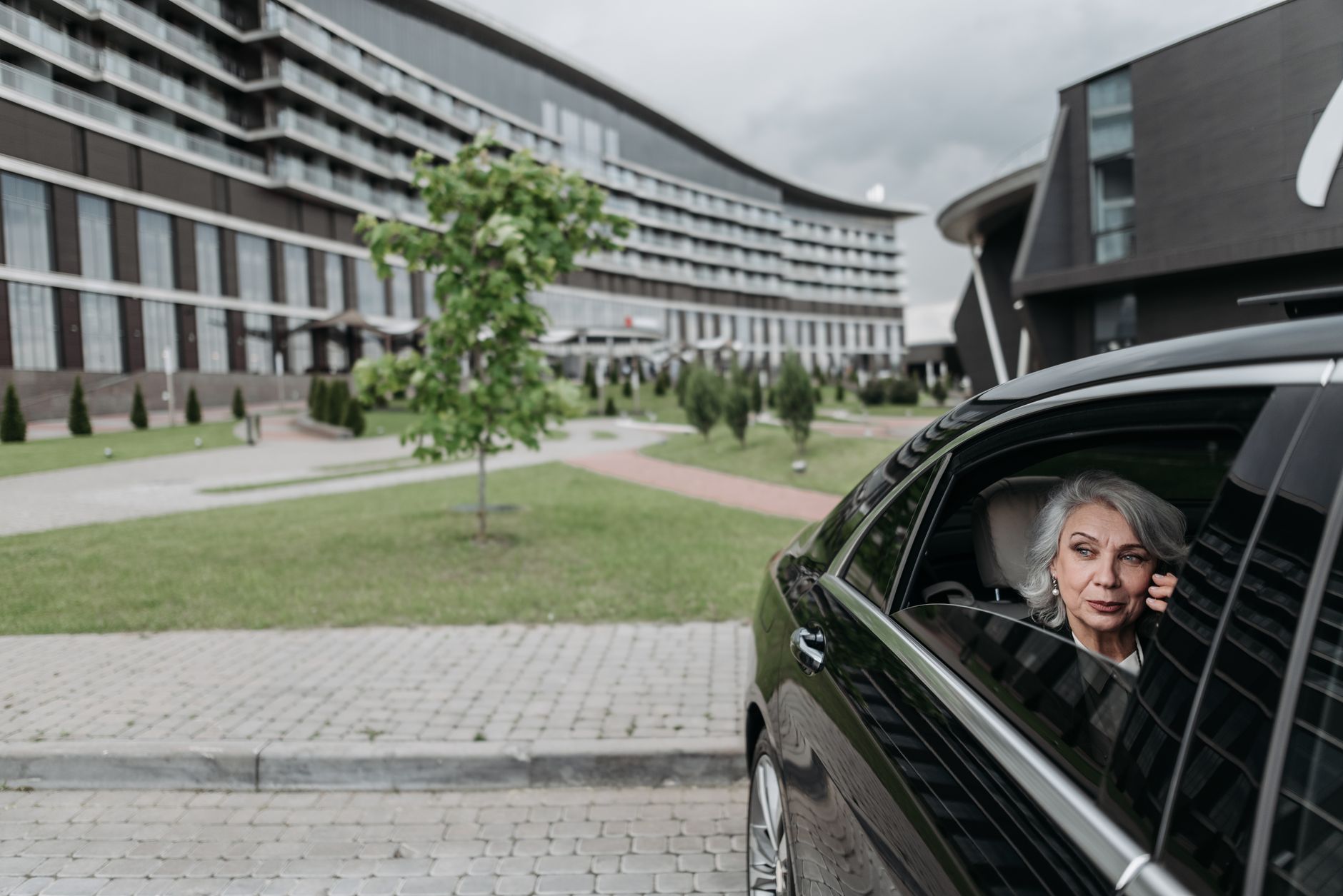 An elderly woman speaks on the phone from a luxury car at a modern clinic.