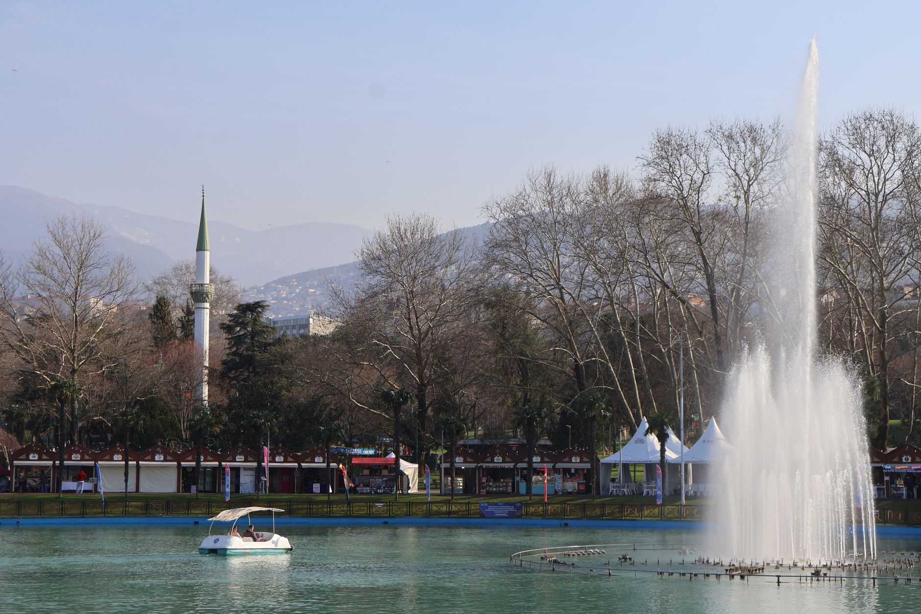 A tranquil view of a lake in Bursa featuring a fountain, a boat, and a mosque in the background.