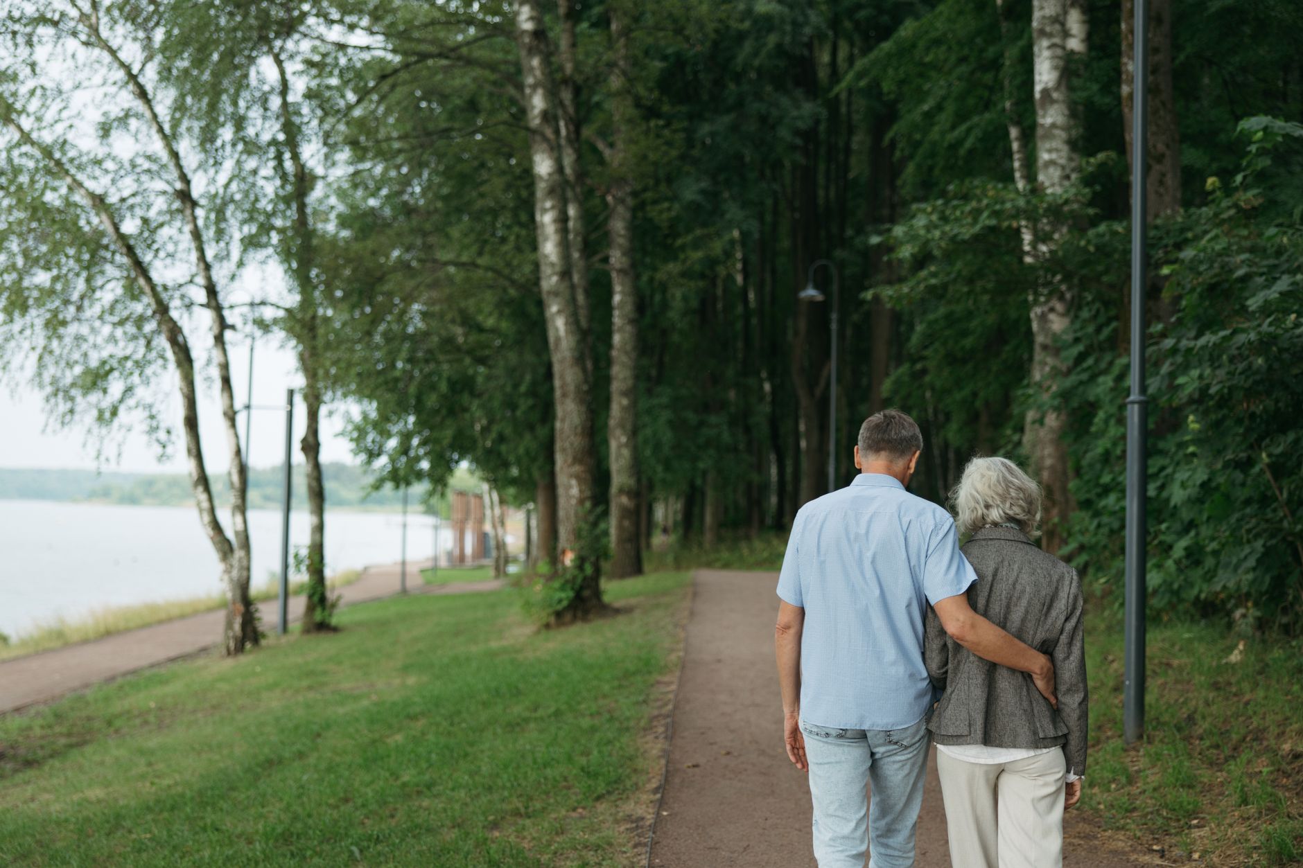 A couple strolls hand in hand along a lakeside path, surrounded by trees.