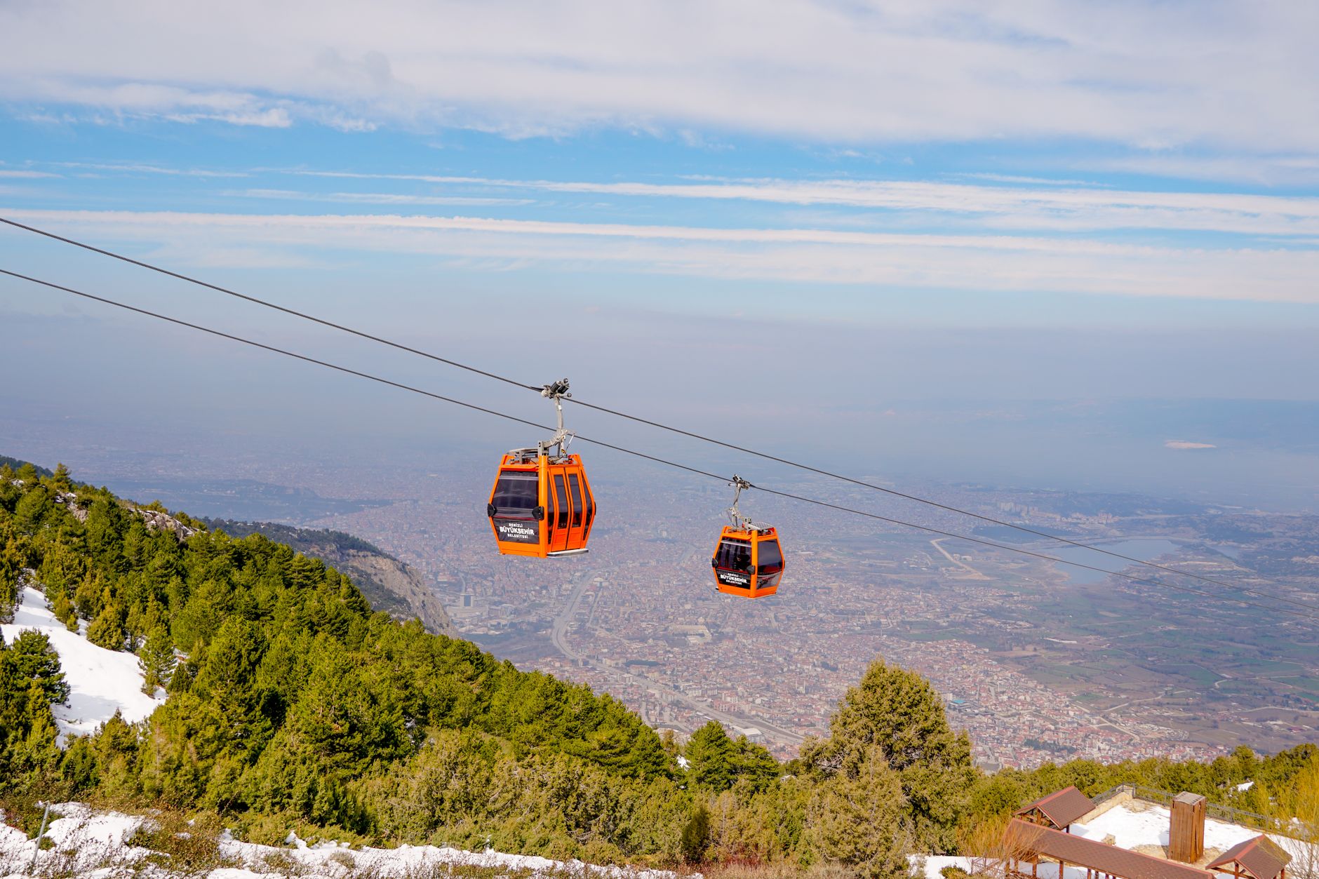 Two orange cable cars traverse the mountains above İzmir, surrounded by greenery and a vast cityscape.