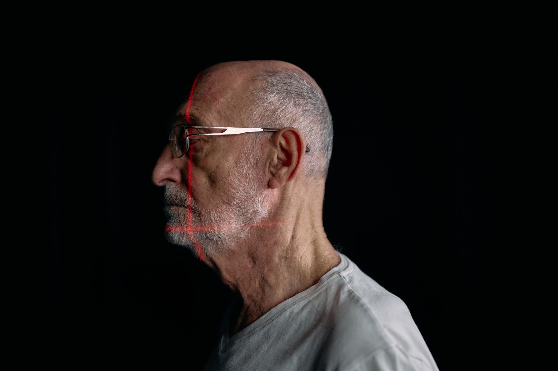 Profile of an older man with glasses in a dark setting, laser lines projected on his face.