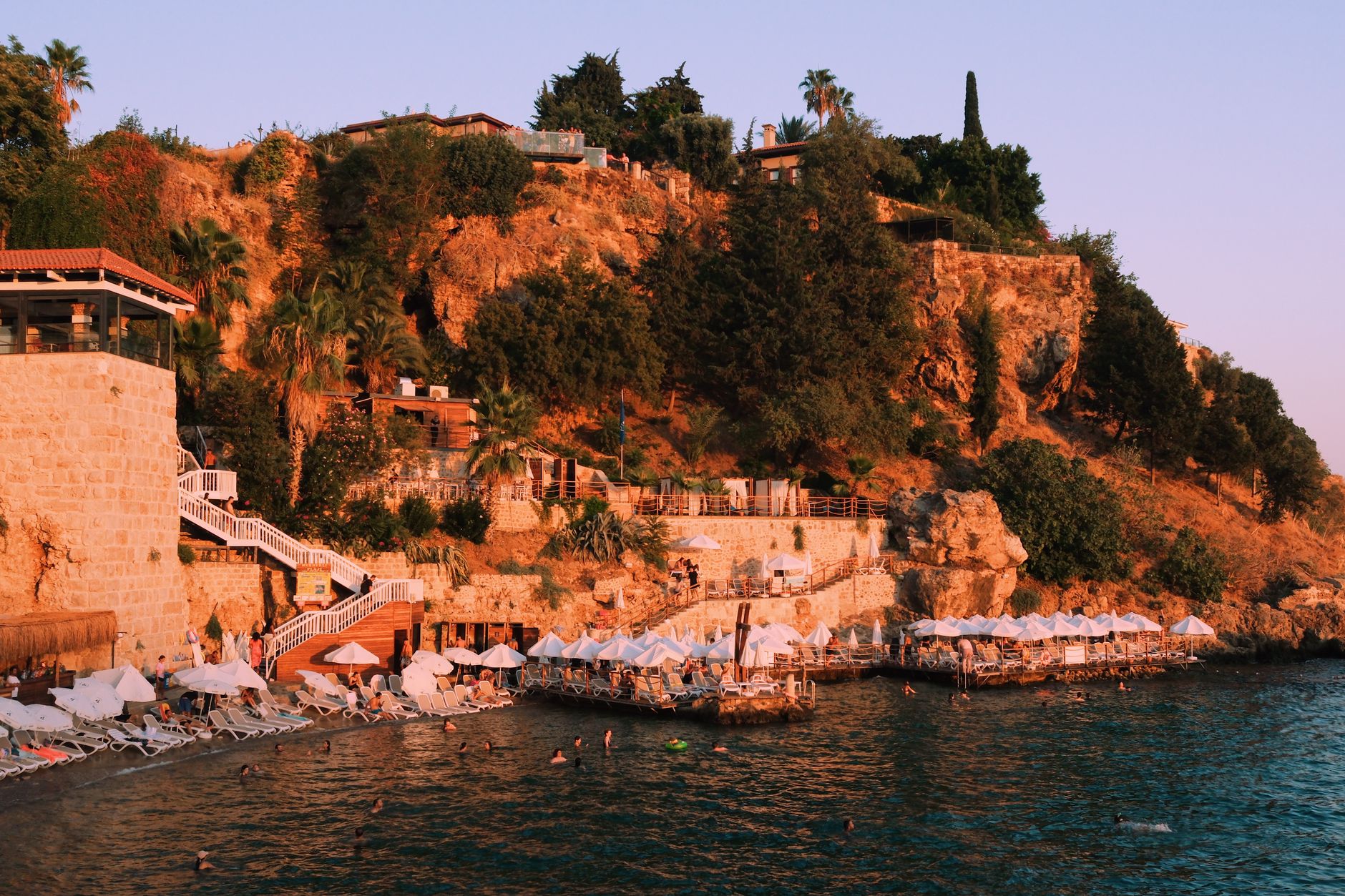 Tranquil Antalya coastline with beach chairs and people swimming at sunset.