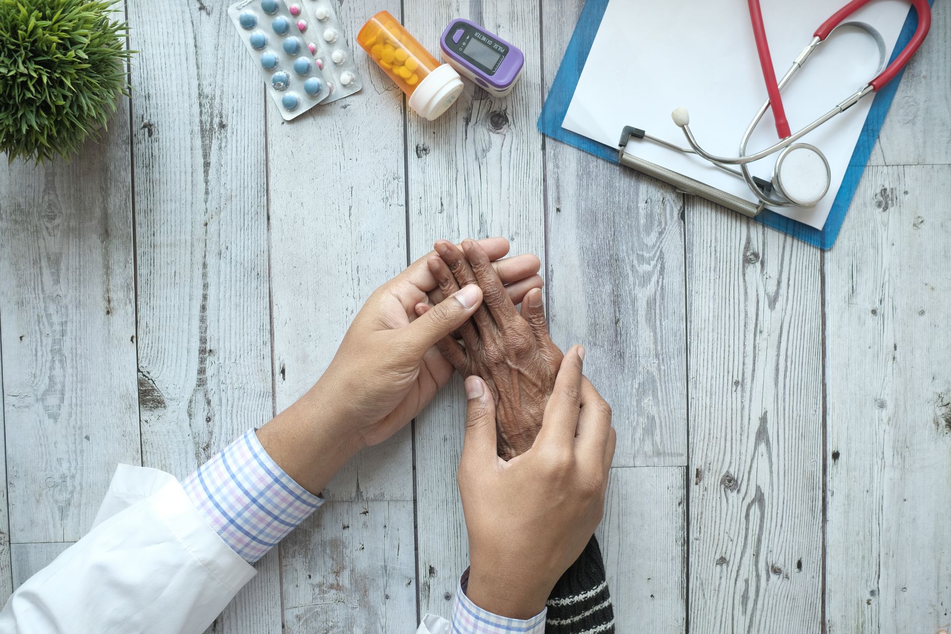A doctor gently holds an elderly patient's hand during a consultation, with medical tools and medications visible.