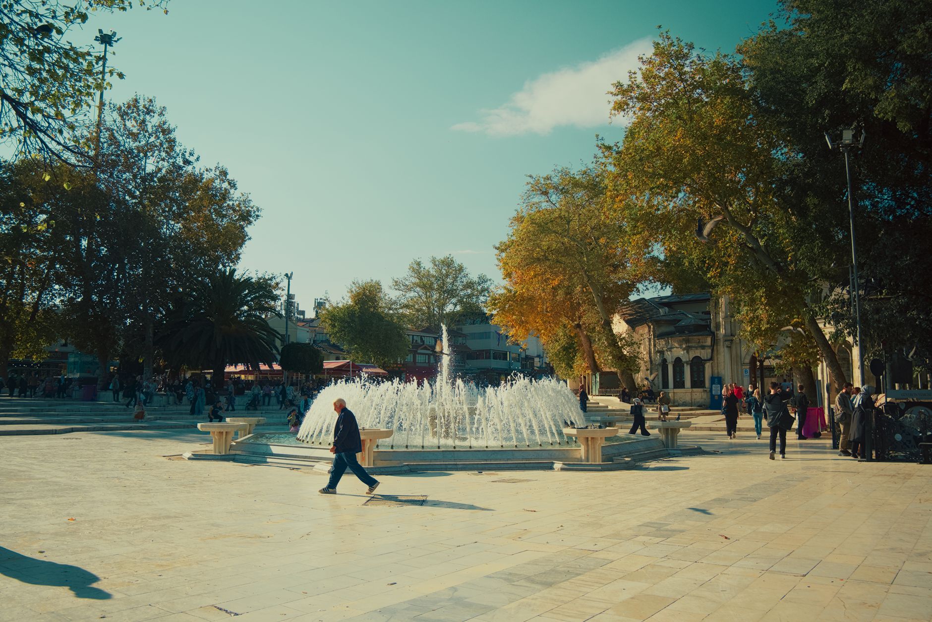 People walking in a sunlit Istanbul park with a fountain and autumn trees.