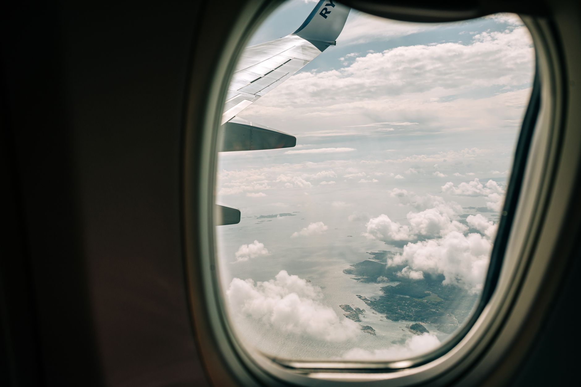 Airplane wing peeking out from window, clouds and distant islands visible over the Gulf, representing flights to Türkiye.