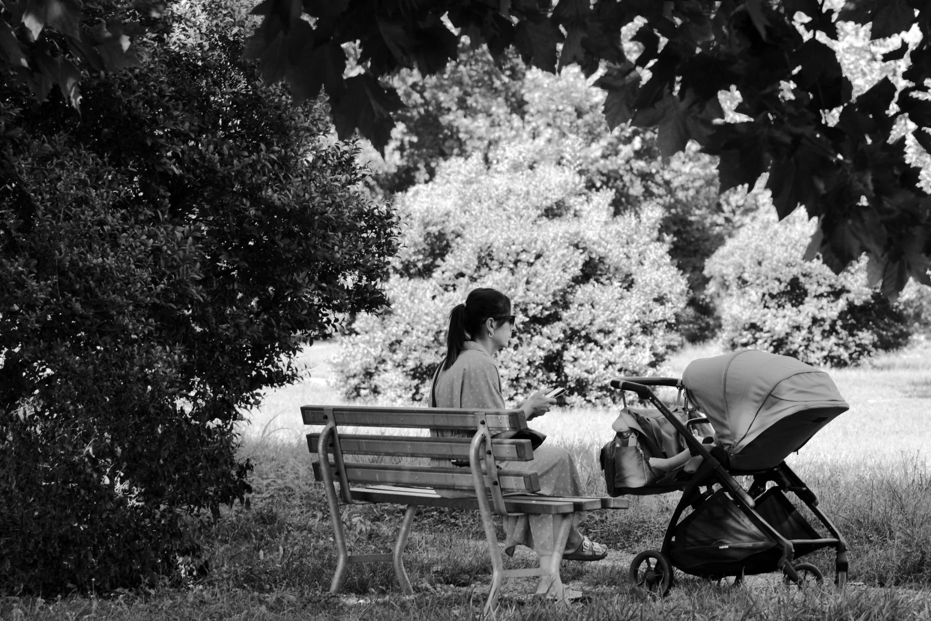 A parent seated on a bench in a park, looking at a phone, with a stroller nearby.