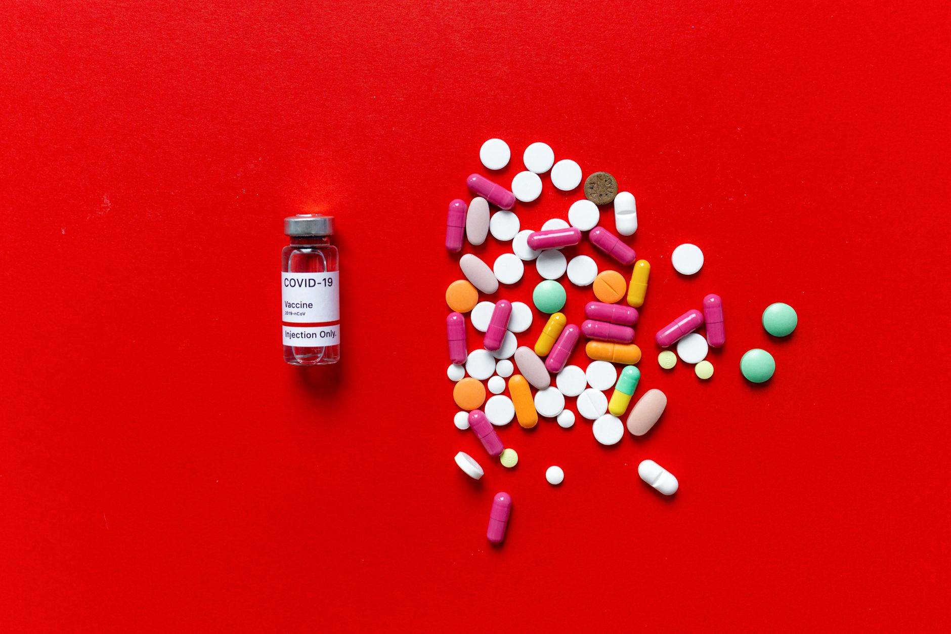 A vial labeled COVID-19 vaccine next to various colorful pills on a red background.