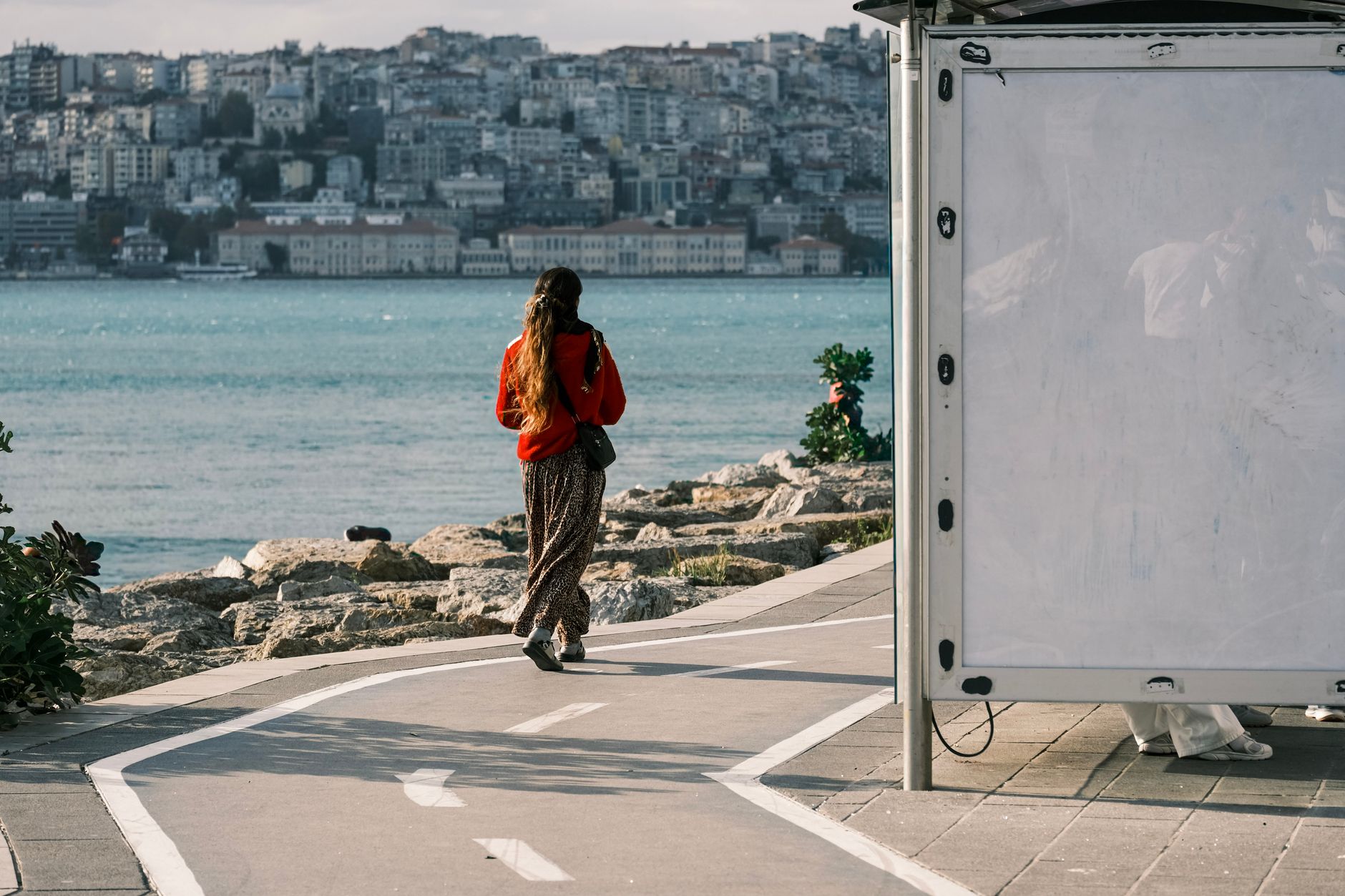 A woman walks on a path by the Bosphorus in Istanbul, with cityscape in the background.