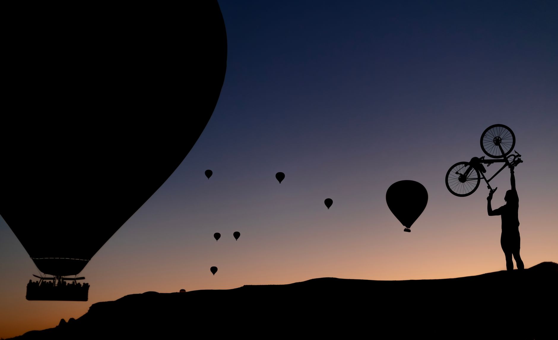 Silhouette of a cyclist holding a bike amidst hot air balloons at sunset.