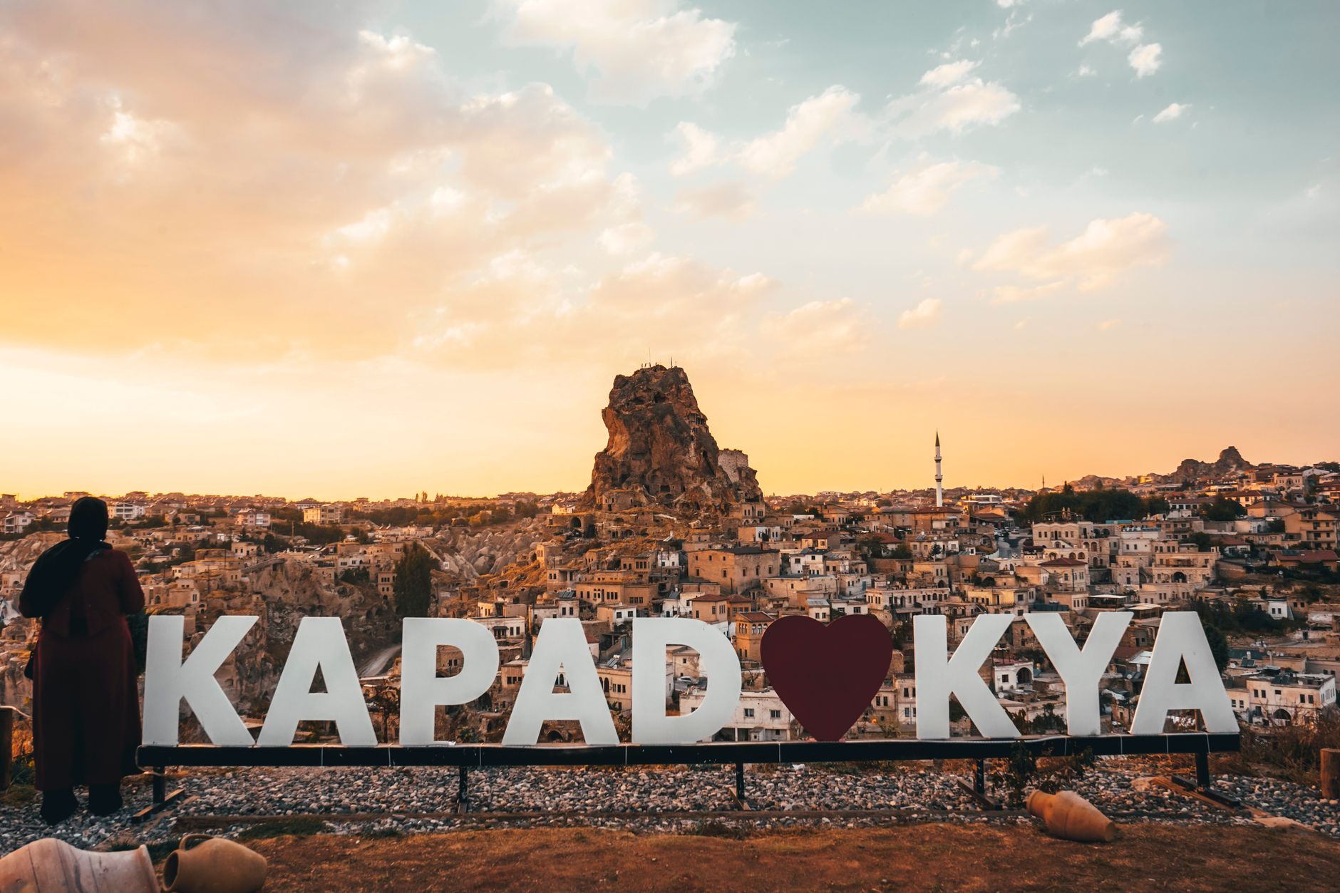 A scenic view of Cappadocia at sunset, featuring a logo and a figure overlooking the landscape.