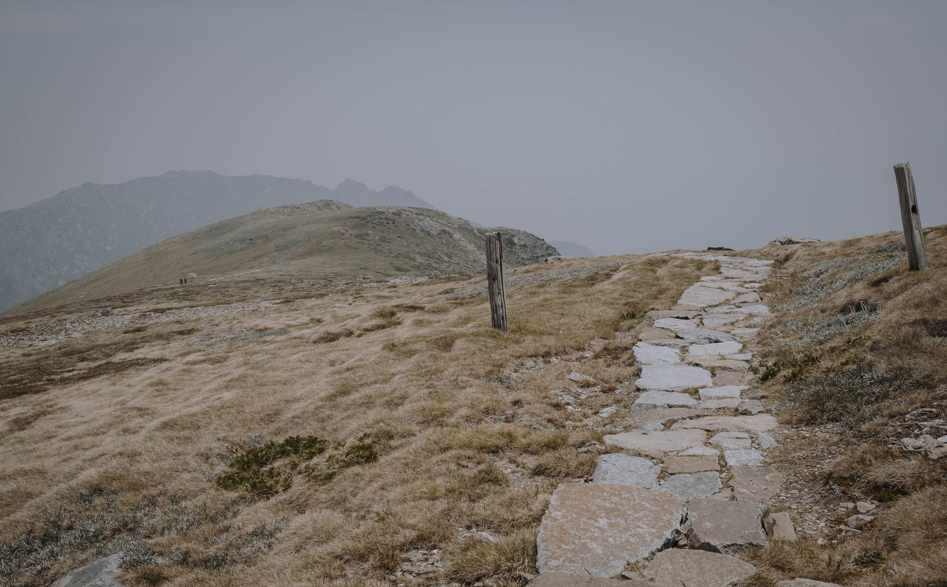 A stone pathway leads across grassy terrain on Mount Honaz, overlooking distant mountains.