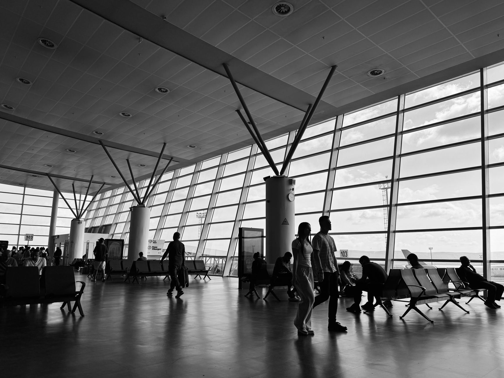 Black and white photo of passengers at Tbilisi airport, waiting by large windows.