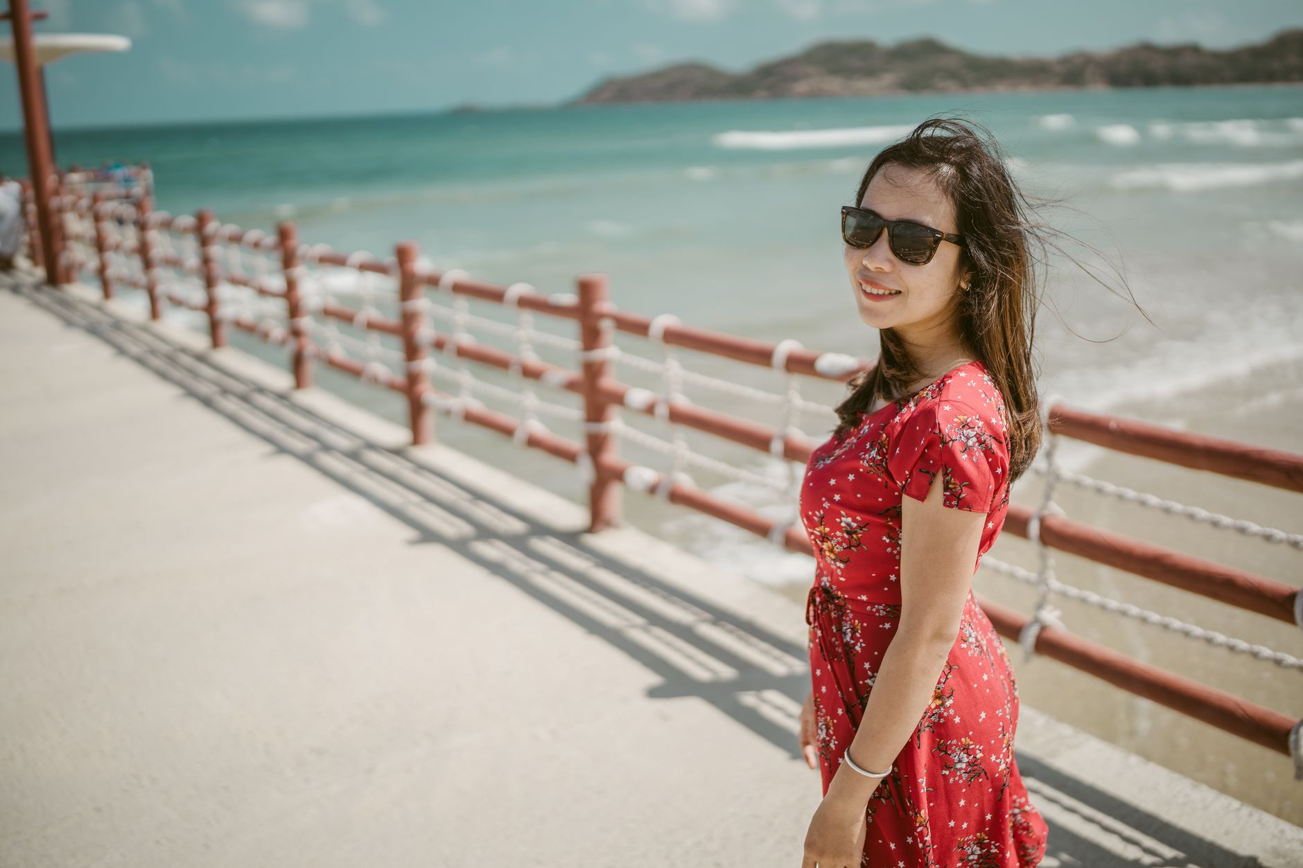 A cheerful individual in a red floral dress enjoys a sunny day at Antalya beach, promoting aligner benefits.